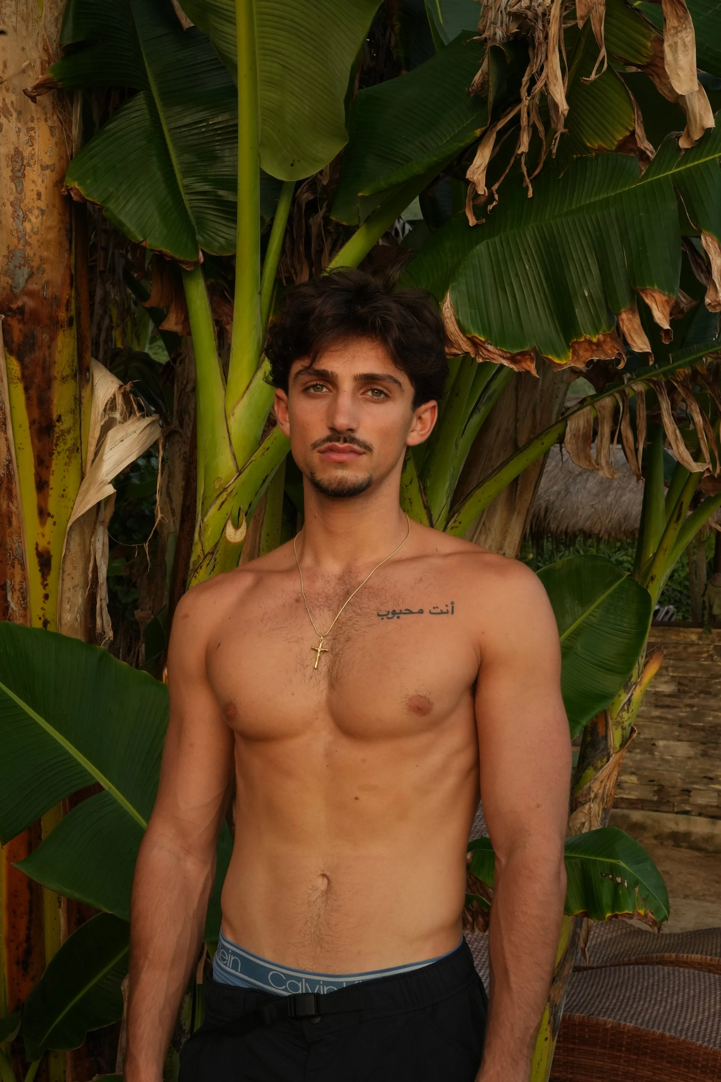 A shirtless young man with dark hair and a goatee standing in front of large green banana leaves, wearing black shorts, a gold cross necklace, and a tattoo in Arabic on his chest.