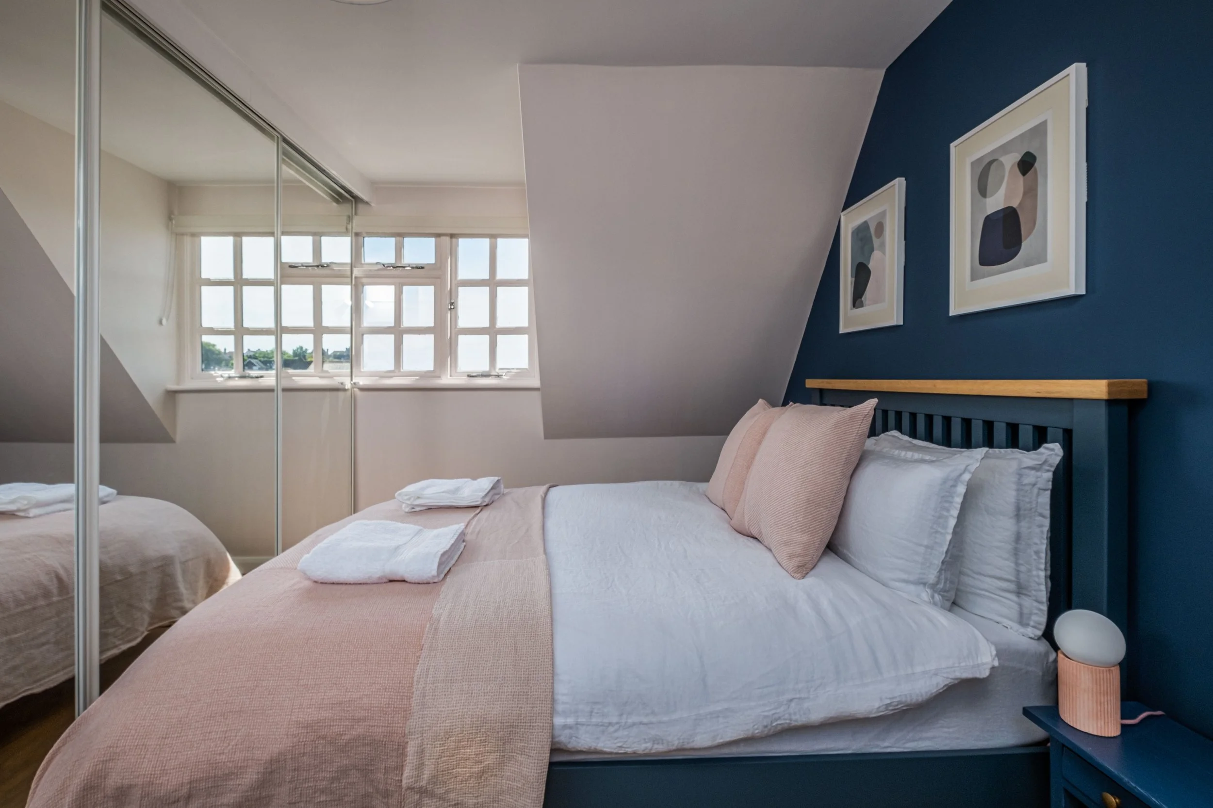 A cozy bedroom with a blue accent wall, framed abstract art, a bed with white and blush bedding, and a window with sunlight streaming in.