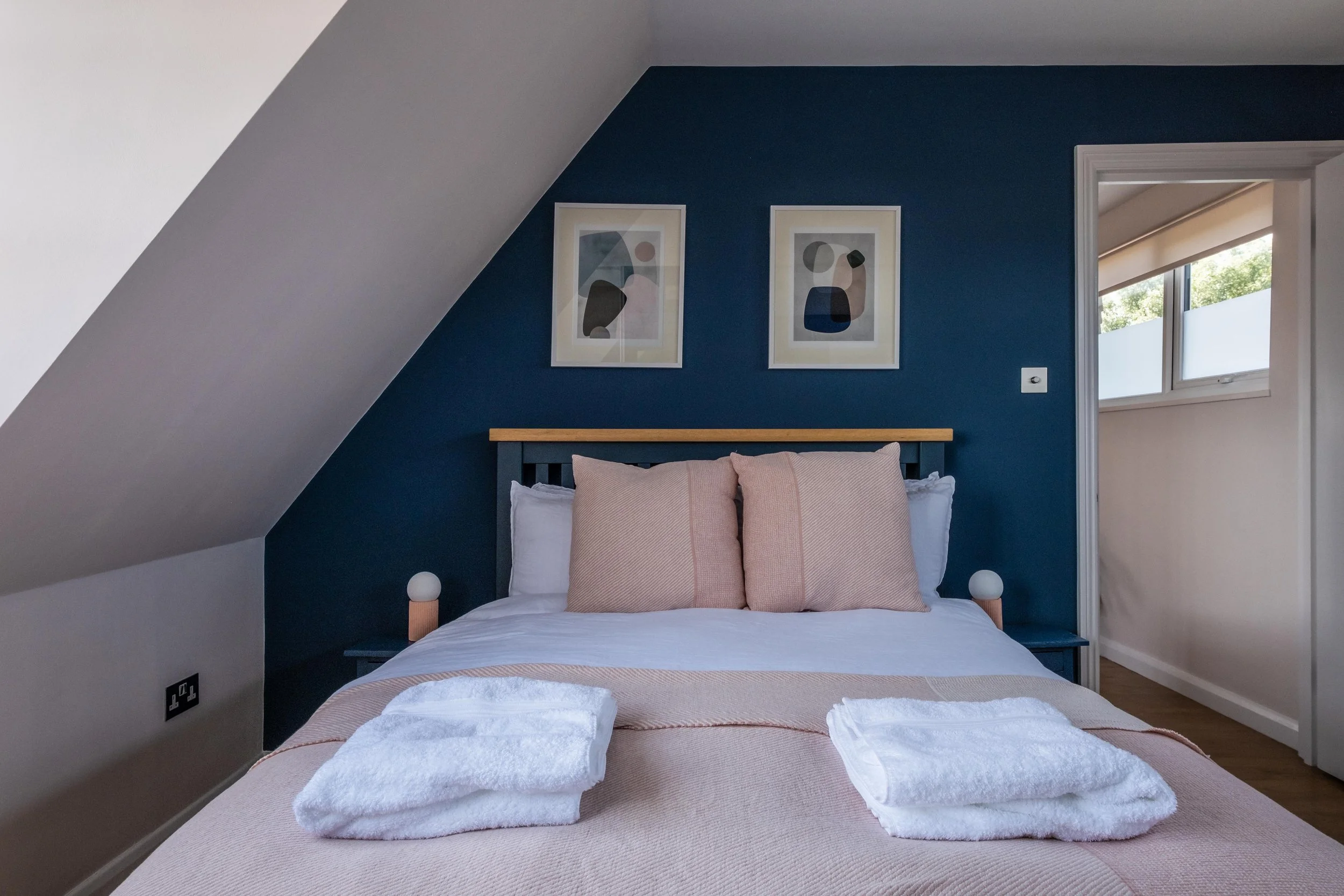 A neatly made bed with light pink pillows and bedspread, towels folded on the bed, against a dark blue accent wall with two framed abstract art prints, a window with sunlight, and a door leading to another room.
