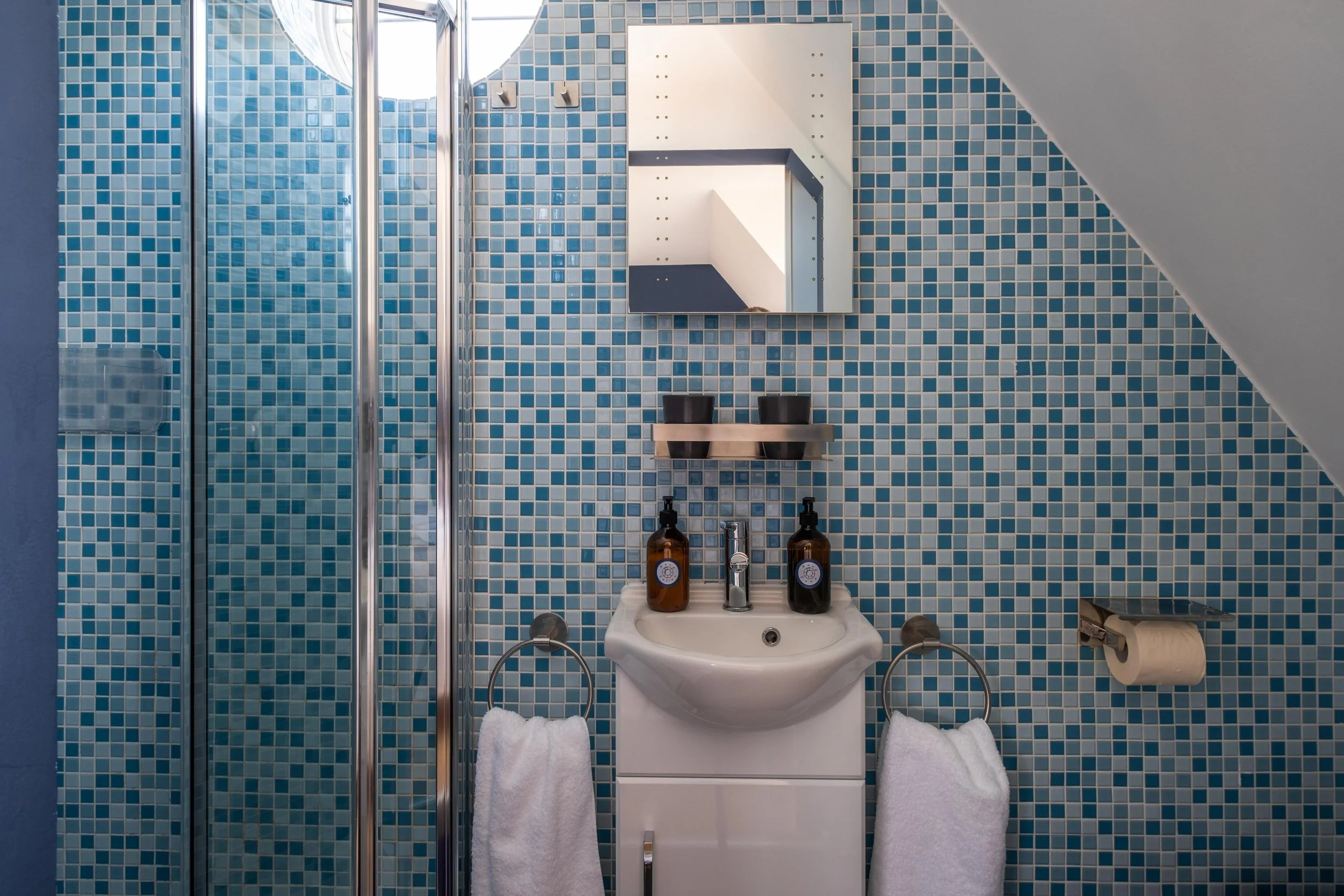 A small bathroom with a blue mosaic tiled wall, a white sink with two brown soap dispensers, a mirror with a shelf holding two black cups, a hand towel on a ring, a toilet paper holder with roll, and a glass shower door with metal trim.