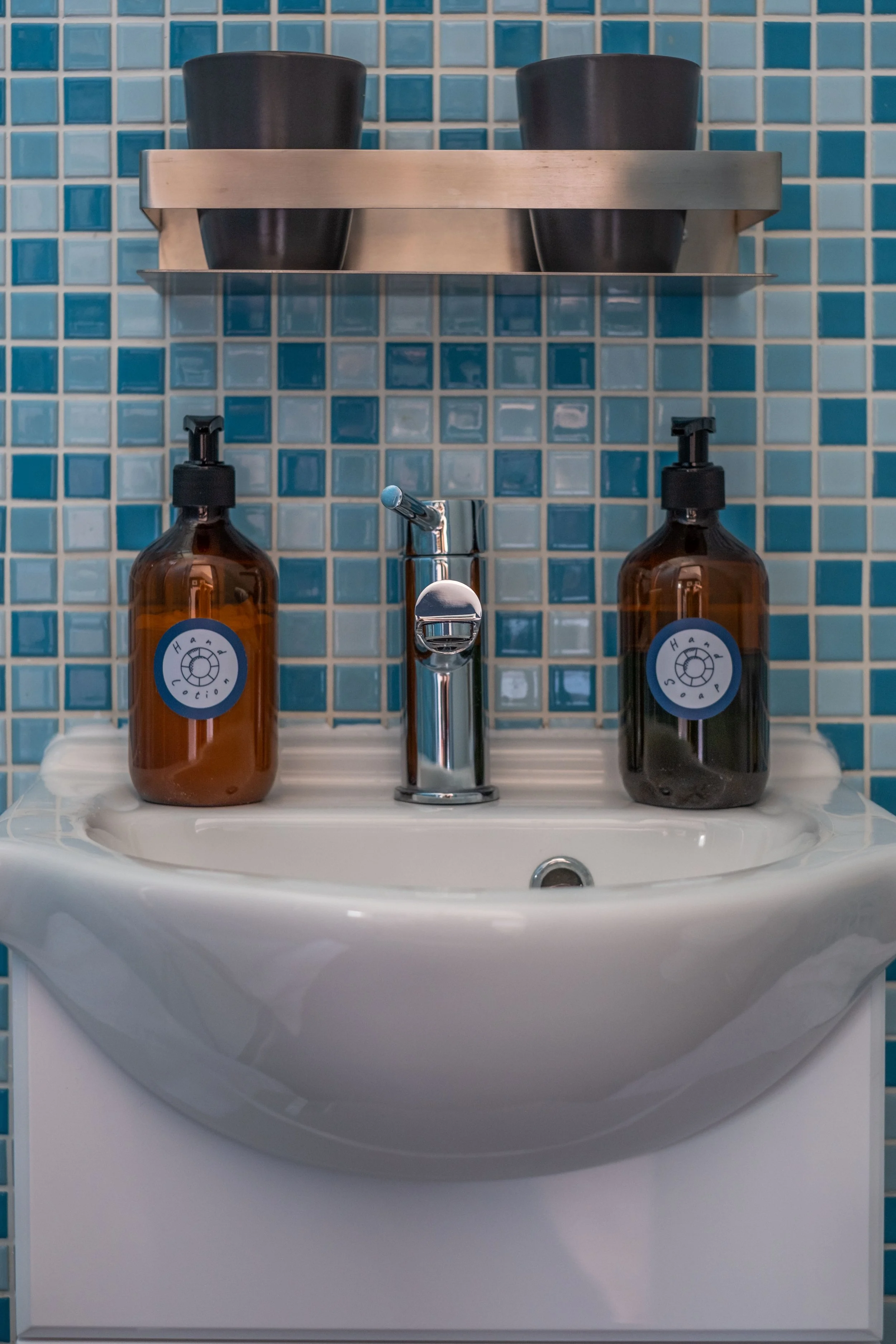 A small white bathroom sink with a silver faucet, two amber-colored soap dispensers labeled 'Hand Soap', mounted on a blue tiled bathroom wall, with a metal shelf holding black cups above the sink.