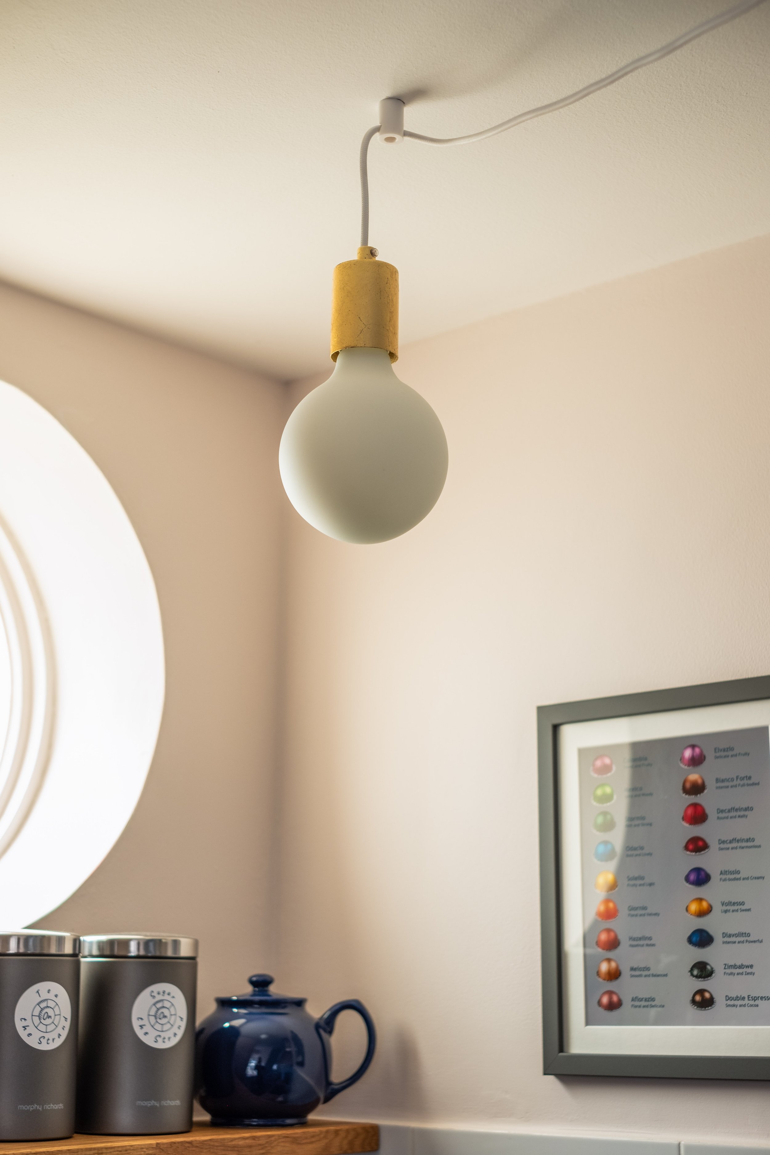 A minimalist lightbulb hanging from the ceiling in a room with a soft-colored wall, a framed coffee color palette on the wall, and containers with labels on a wooden shelf.