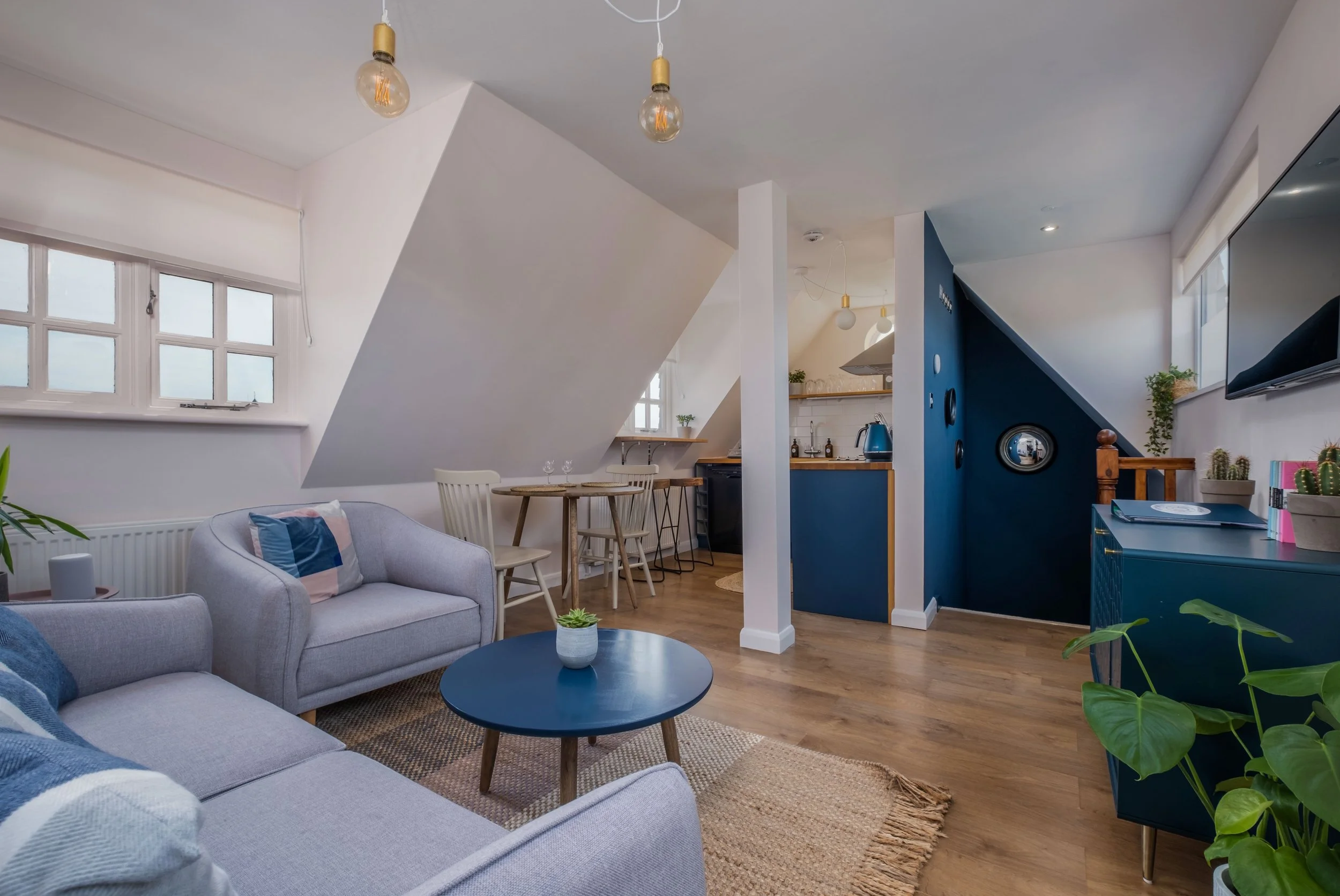 Living room with gray couches, a black coffee table, and green plants, opening to a small dining area and kitchen with white cabinets and blue walls, under sloped ceilings with hanging light bulbs.