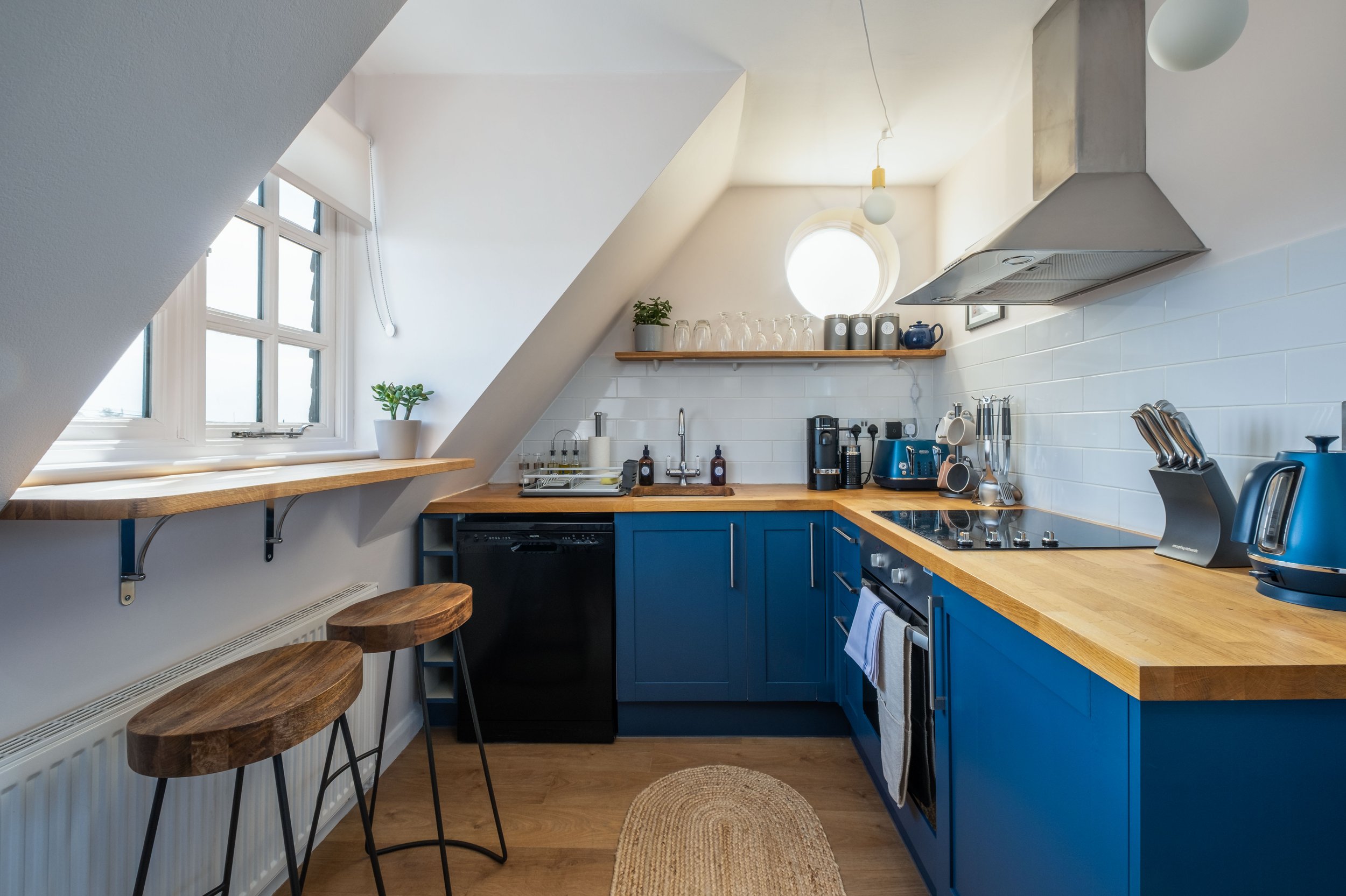 Small modern kitchen with blue cabinets, wooden countertops, white tiled backsplash, and angled ceiling. Features a window with a wooden shelf, two barstools, and various appliances including a coffee maker, kettle, and stove.
