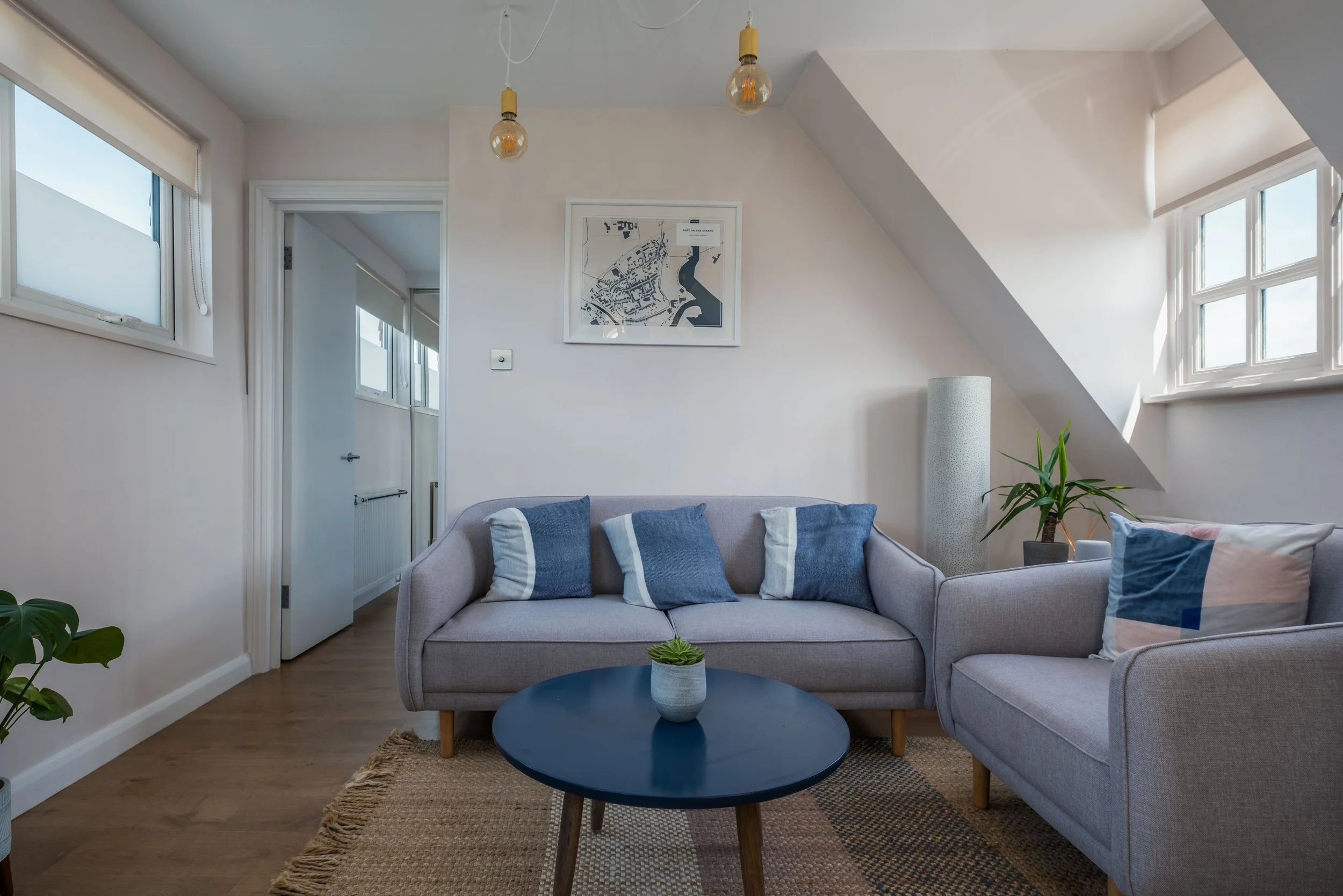 Living room with two light gray sofas, blue and white cushions, a dark blue coffee table with a small plant, a tall white floor lamp, and potted plants near windows with natural light.