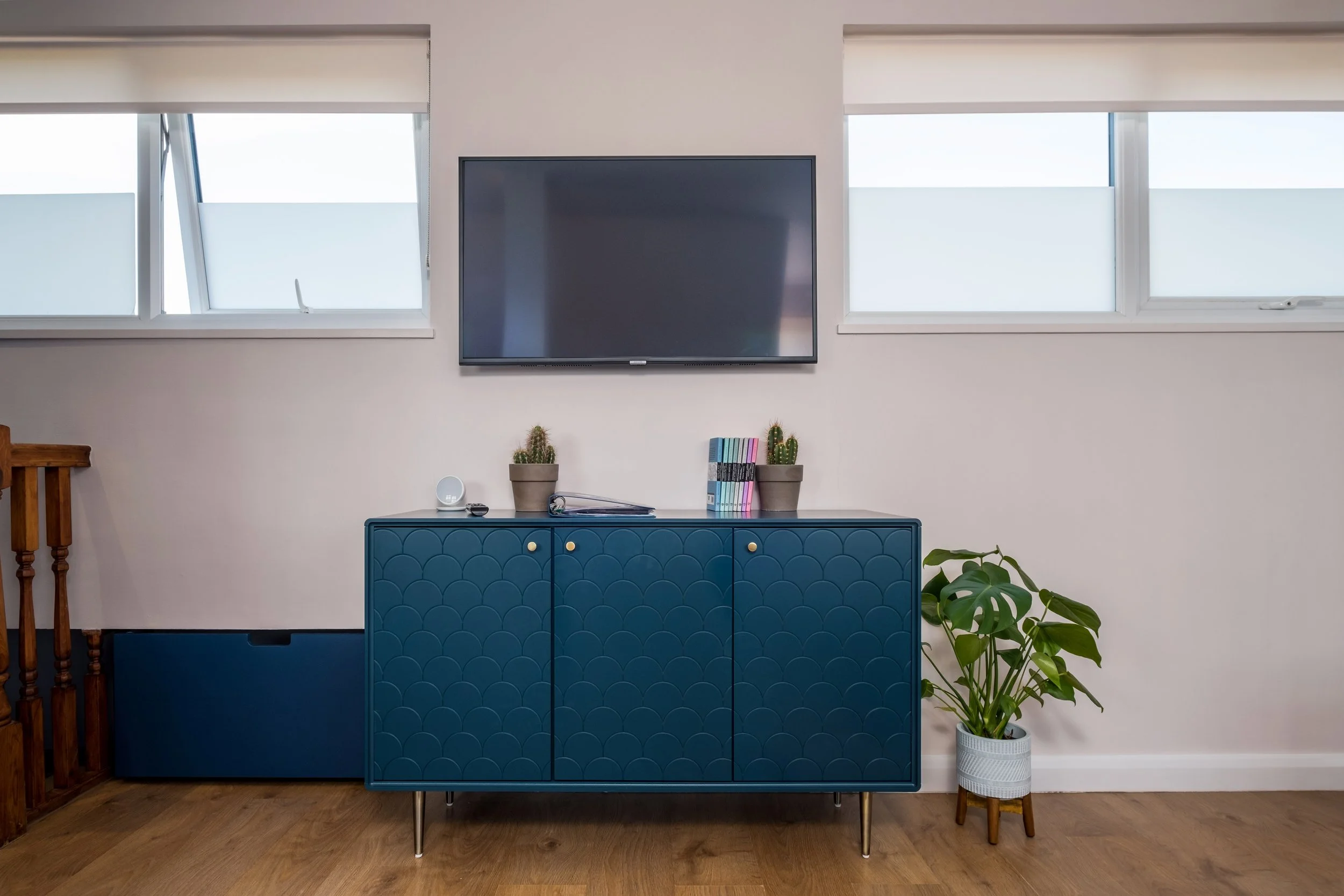 A modern living room scene with a blue cabinet, potted plants, a television, and two windows.