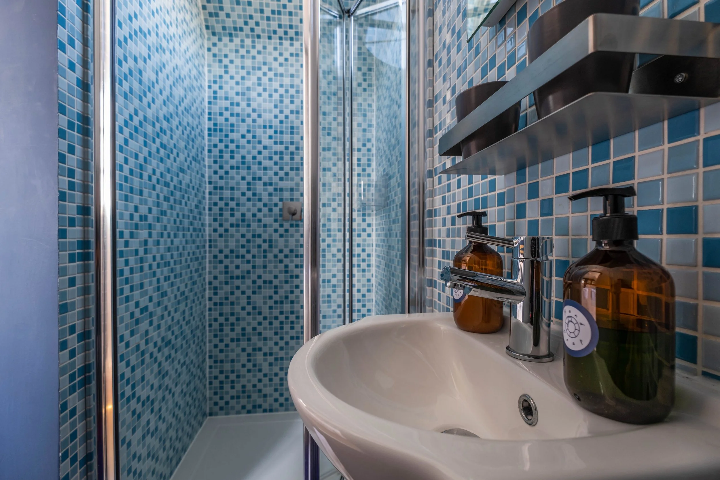 Small bathroom with a mosaic tile shower, white sink with a chrome faucet, and amber soap dispensers on the sink counter.