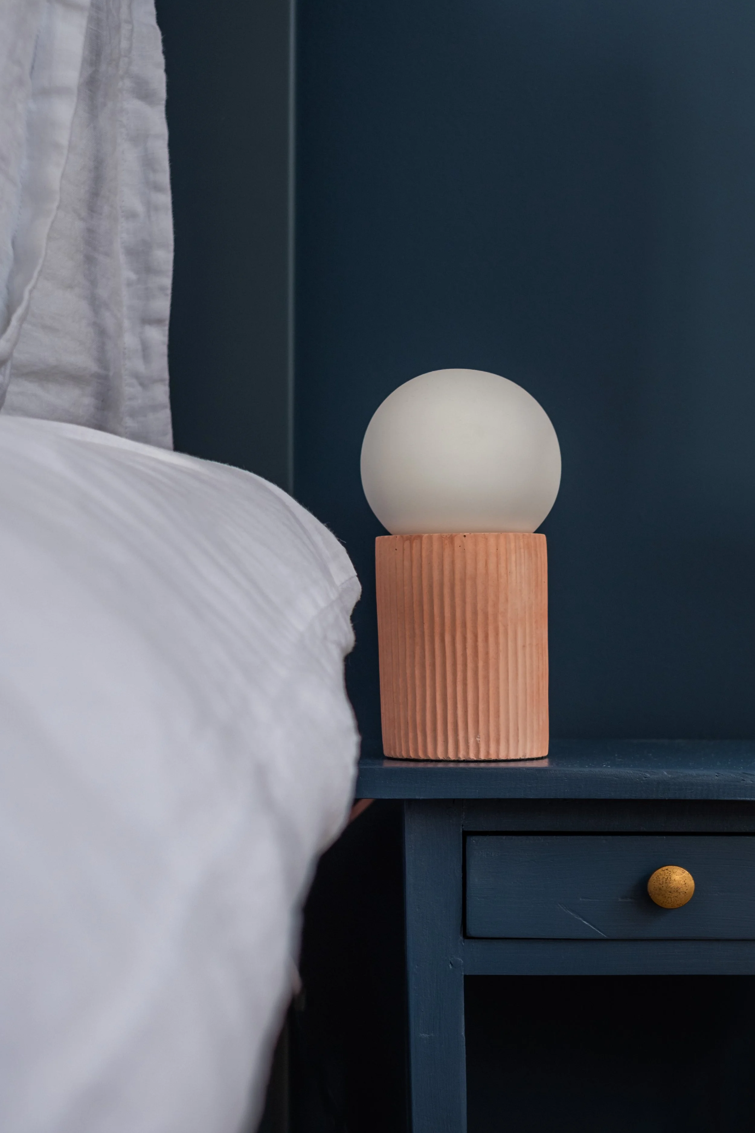 Close-up of a modern bedside table with a round lamp on top, next to a bed with white bedding, against a dark blue wall.