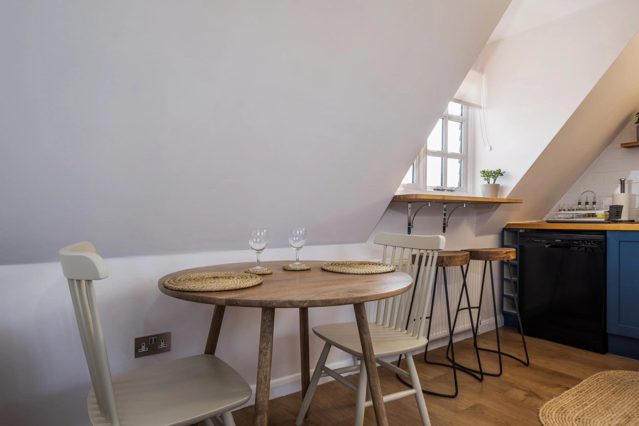 A cozy kitchen area with a wooden dining table, two glasses of water, and a mix of chairs, under a sloped ceiling with window. Includes kitchen counter with potted plant, stove, and shelf.