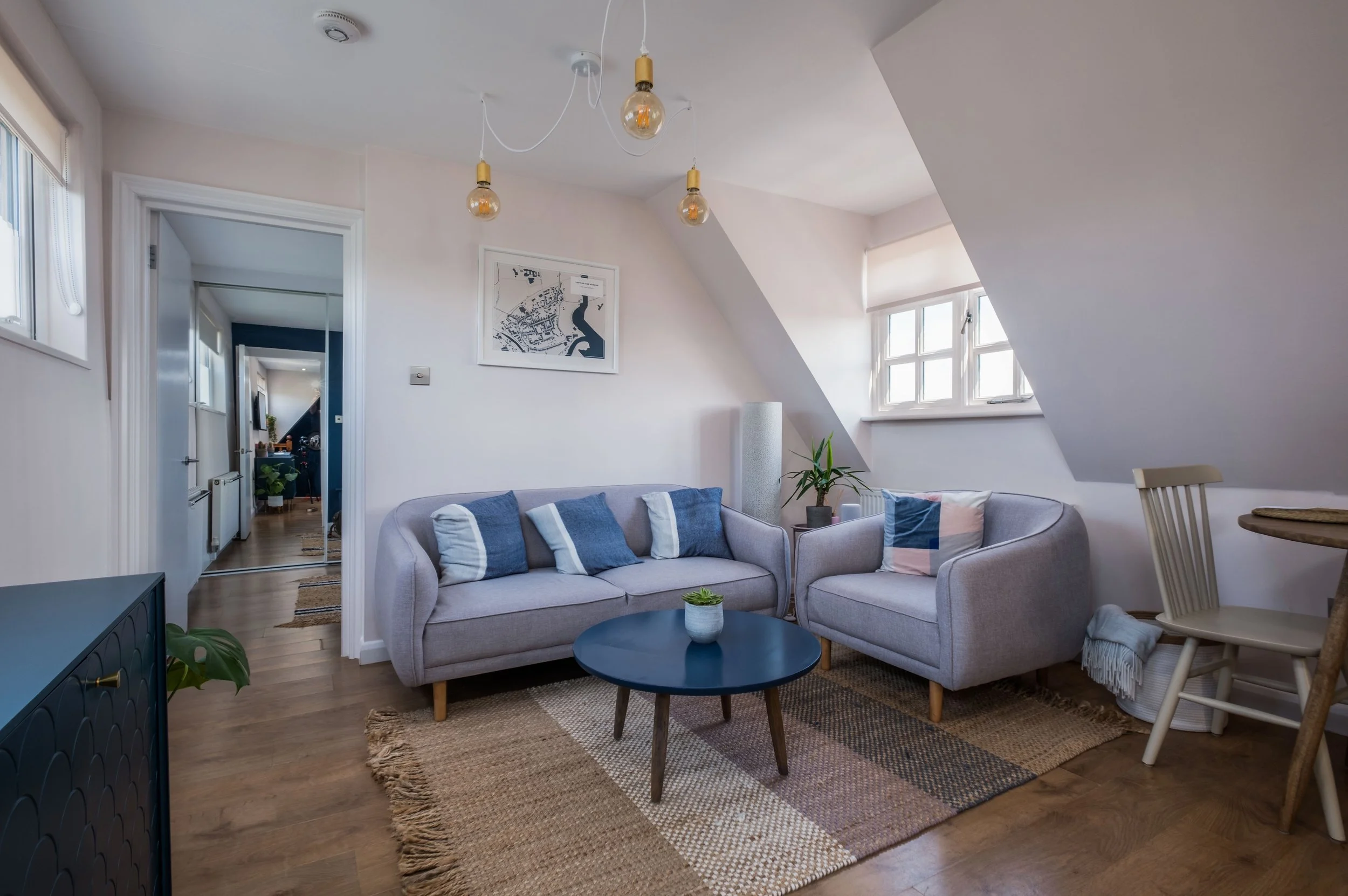 Living room with two light gray sofas, pillows, a black round coffee table, a potted plant, a rug, and white walls with windows and ceiling lighting.