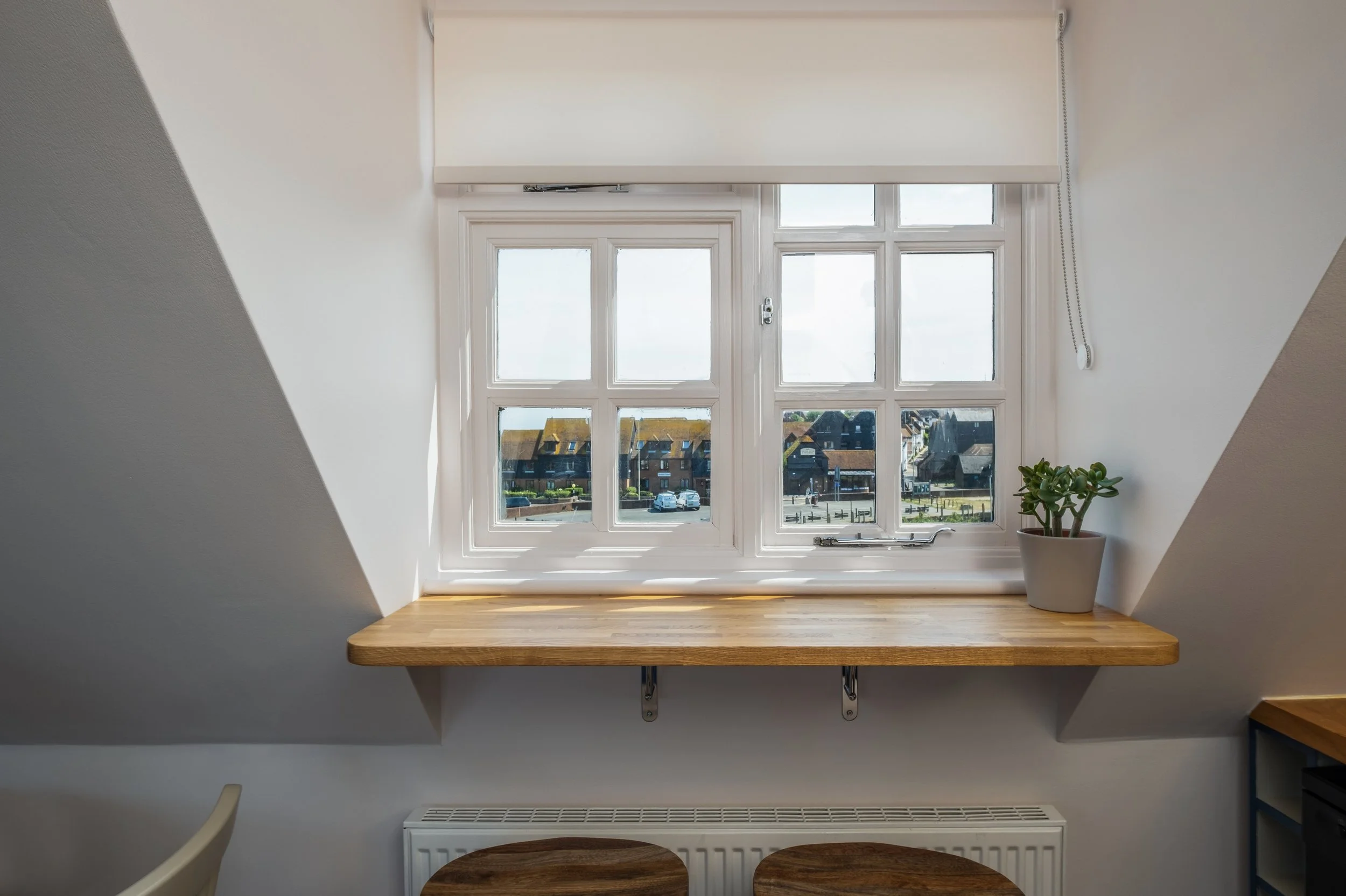 A view from inside a room looking out a white-framed window with multiple panes, a wooden sill, and a white roller blind. There is a small potted succulent on the right side of the sill, and the scene outside shows houses and parked cars.
