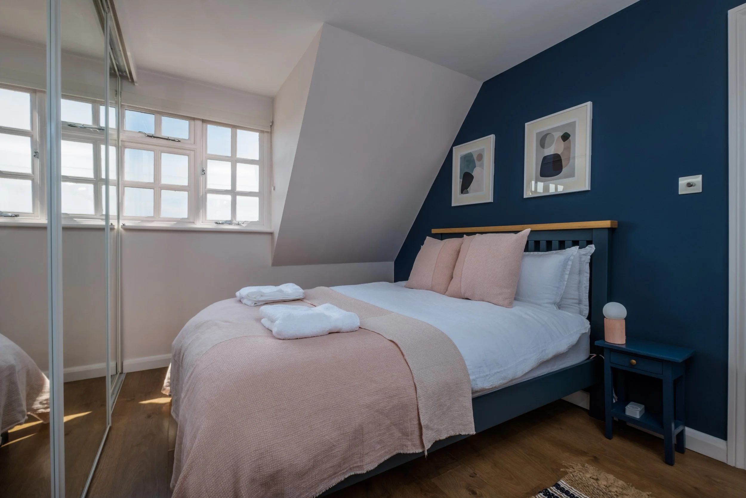 Sunlit bedroom with a bed, pink bedding, beige pillows, navy blue wall, framed abstract artwork, small navy blue nightstand with a modern lamp, and mirrored closet doors.