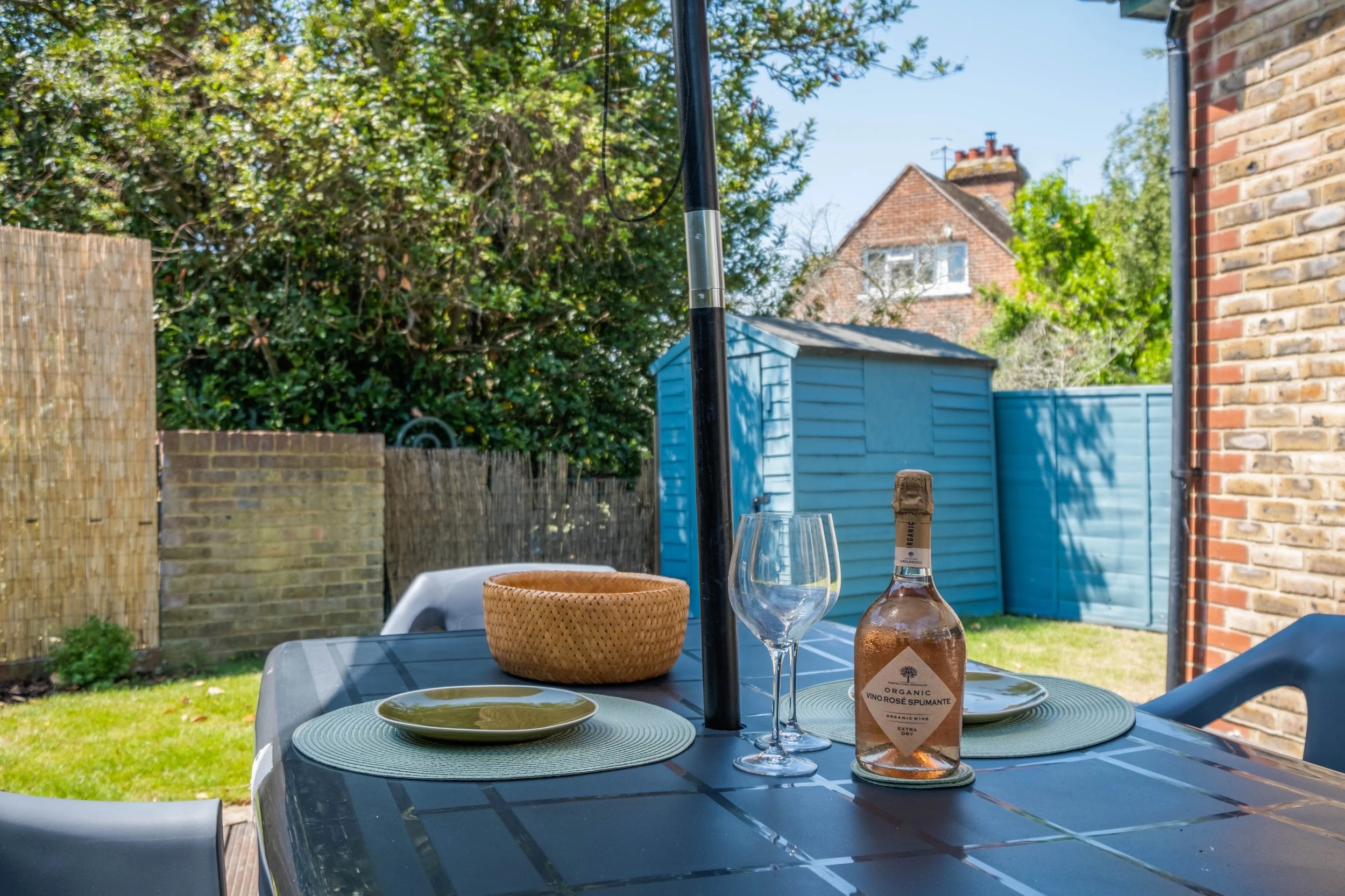 Outdoor patio table with a bottle of sparkling wine, empty wine glass, two plates with green napkins, a woven basket, and a black umbrella, set in a backyard with a green lawn, trees, and blue storage sheds.