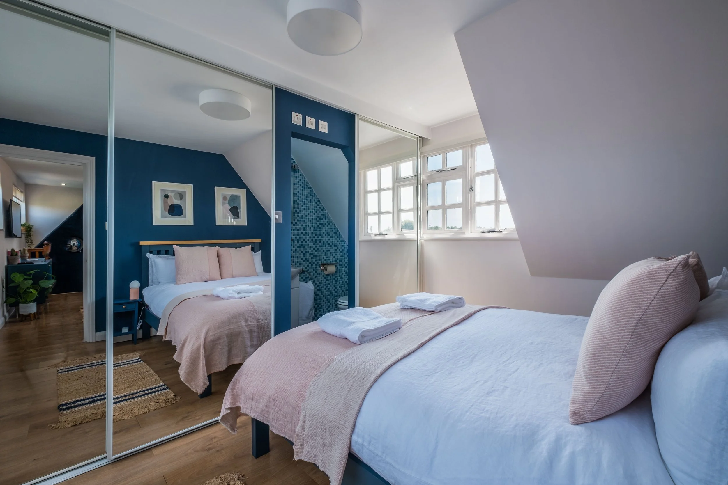 Bright bedroom with a large bed, pink bedding, and fluffy pillows, adjacent to a small bed with pink bedding and artwork on the blue wall, a window with natural light, and a bathroom with blue tiled wall visible through an open partition.