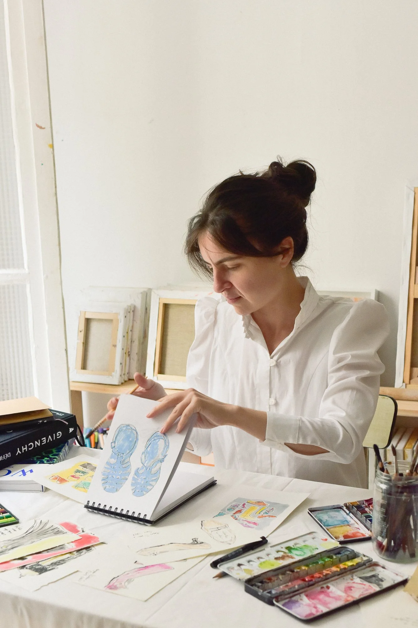 A woman with dark hair tied up in a bun wearing a white shirt, working on a drawing of sneakers in an art studio or classroom. The table is filled with art supplies, including watercolor paints, sketches, and drawings, with framed artwork in the background.