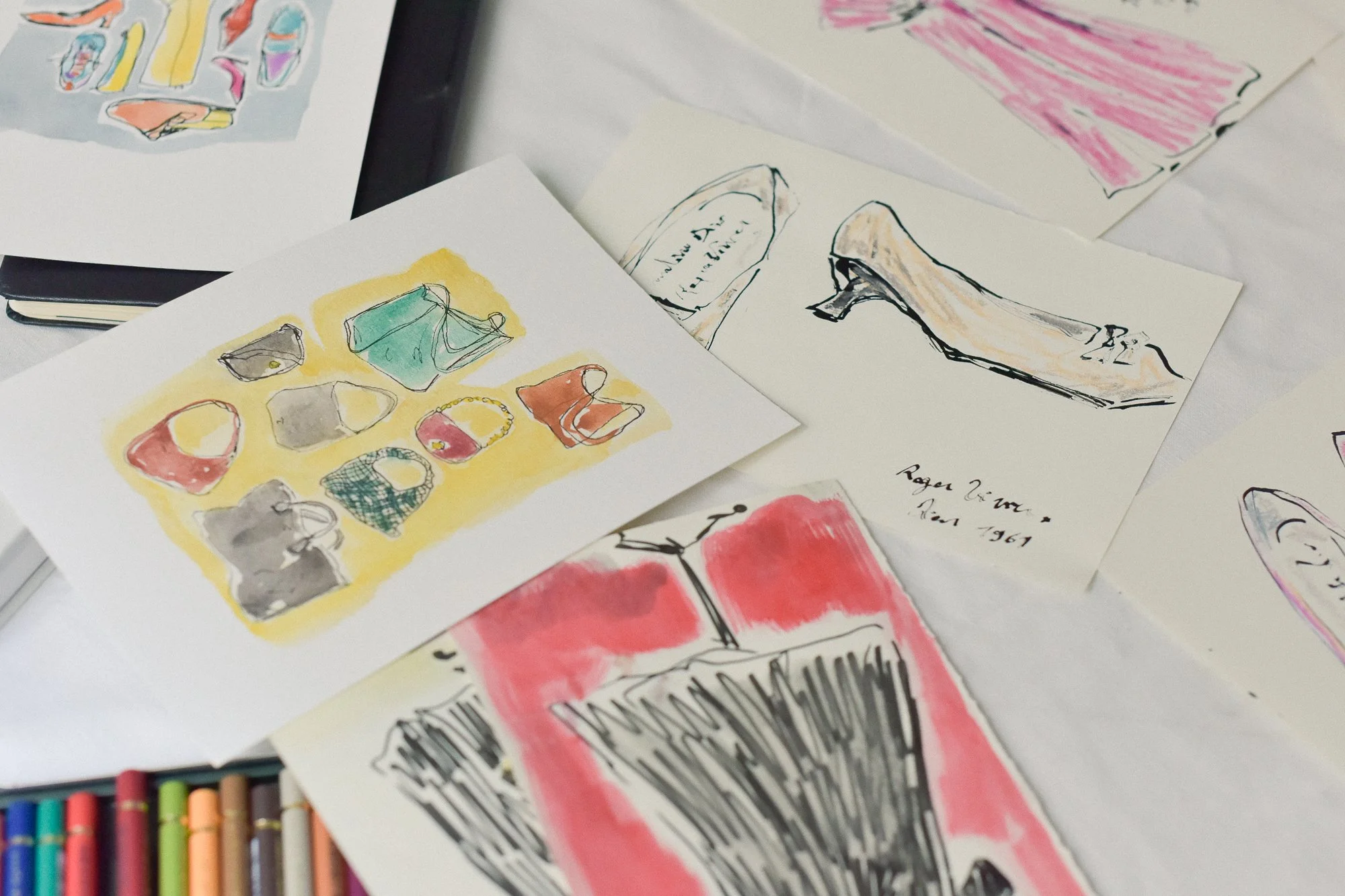 Various colorful watercolor and ink sketches of fashion accessories and shoes on paper, displayed on a white surface with colored chalks at the bottom of the image.