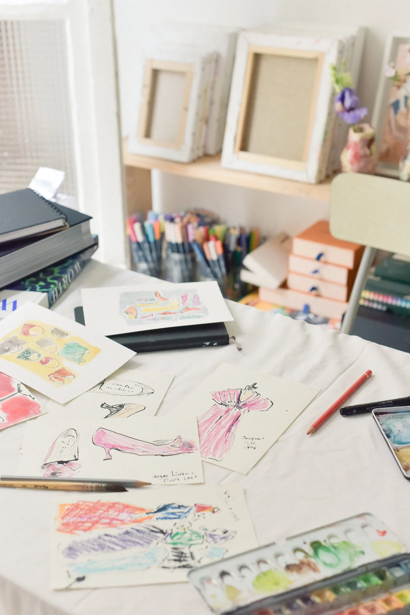 Art supplies and sketches spread out on a table, with some colorful drawings of dresses and other fashion designs, and in the background, framed artworks and art markers.