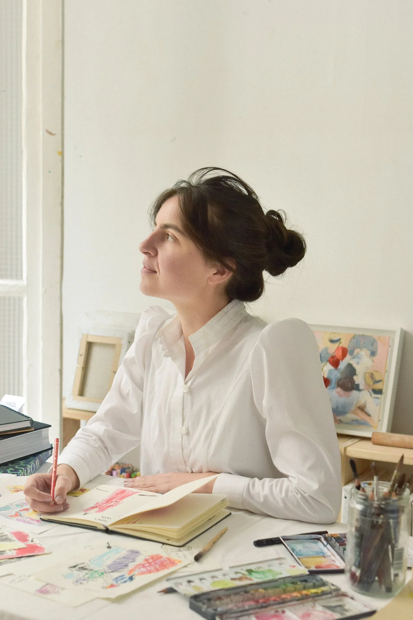 A woman with dark hair styled in a bun, wearing a white blouse, sitting at a desk with art supplies, drawing and coloring on paper.