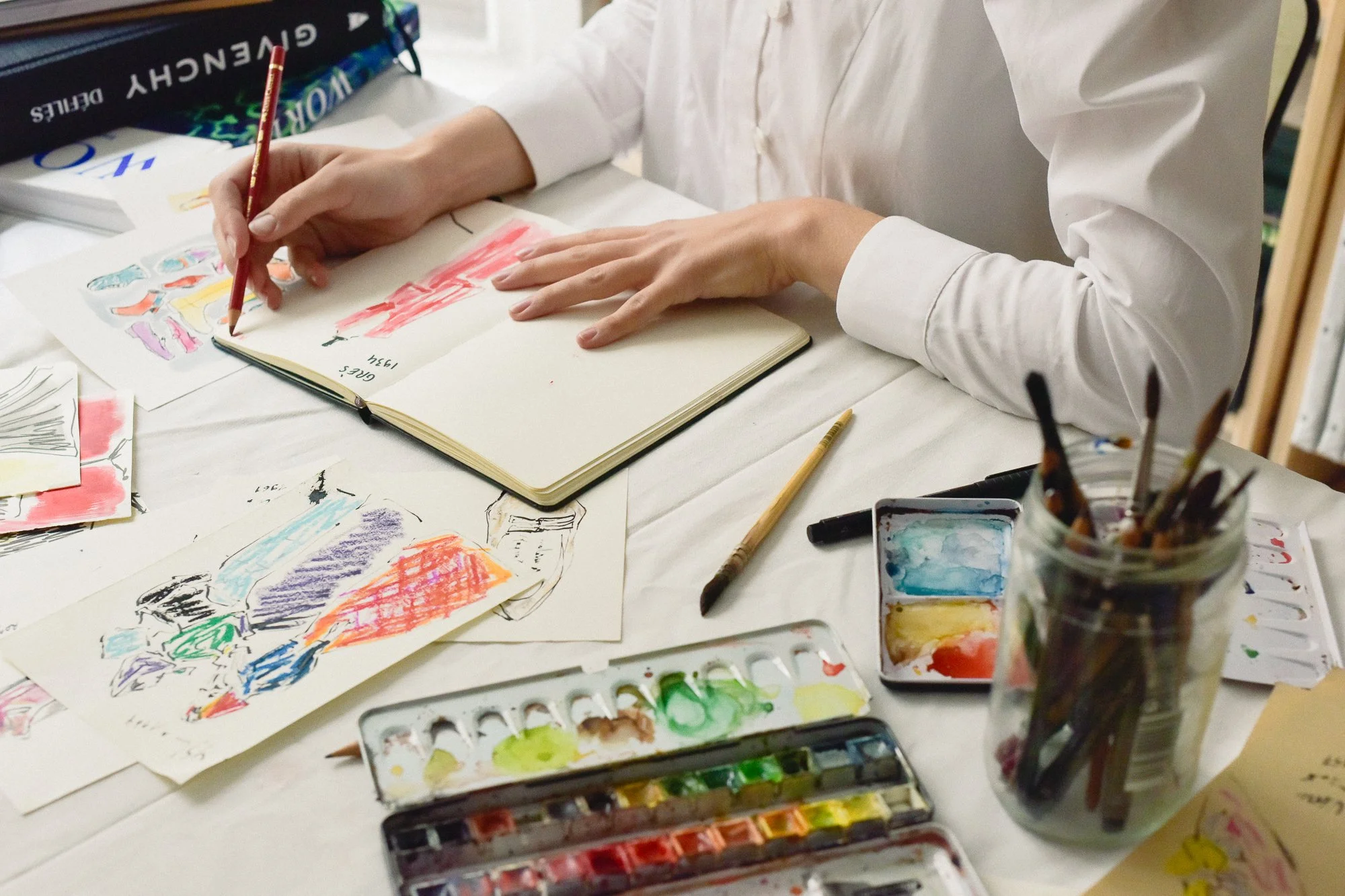Table with art supplies, including watercolor paints, brushes, and sketches. Someone is coloring in a sketchbook with a red pencil. Various colorful drawings and sketches are scattered on the table.