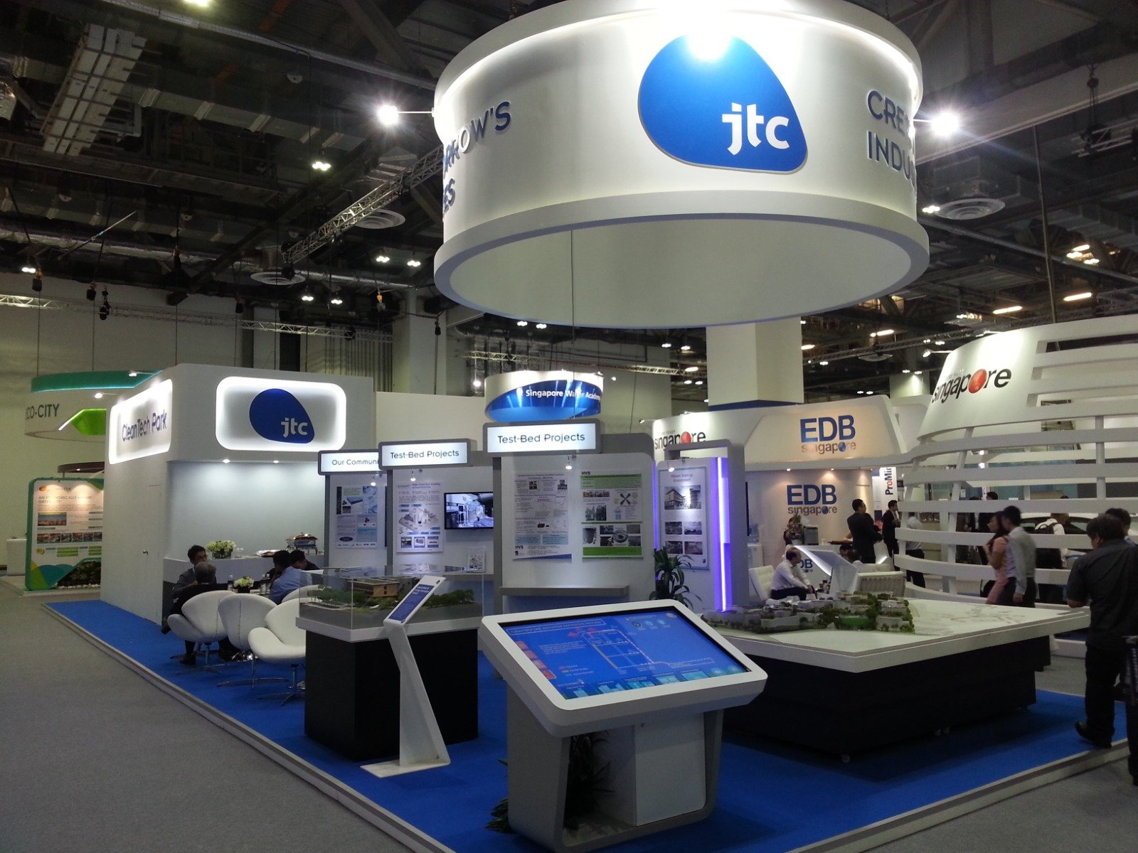 Trade show booth for JTC with displays of projects and information, people walking and viewing exhibits in a large conference hall.