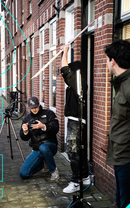 Vrienden maken een fotoshoot buiten een gebouw met rode bakstenen, enkele statieven en een jongen die een camera vasthoudt, terwijl een ander een reflector vasthoudt.