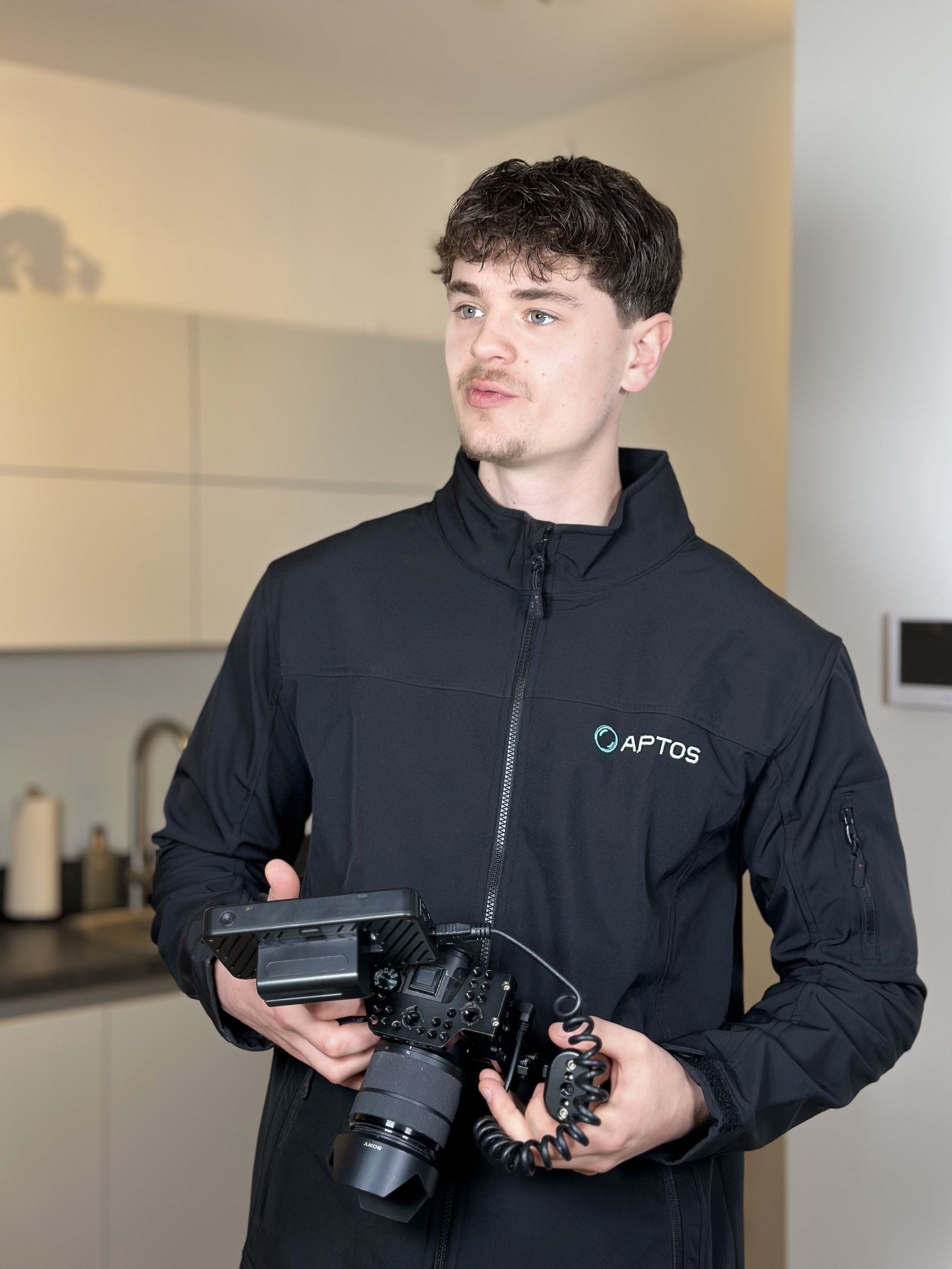 Een jonge man in een zwart Aptos-jack houdt een professionele videocamera in zijn handen in een keuken.