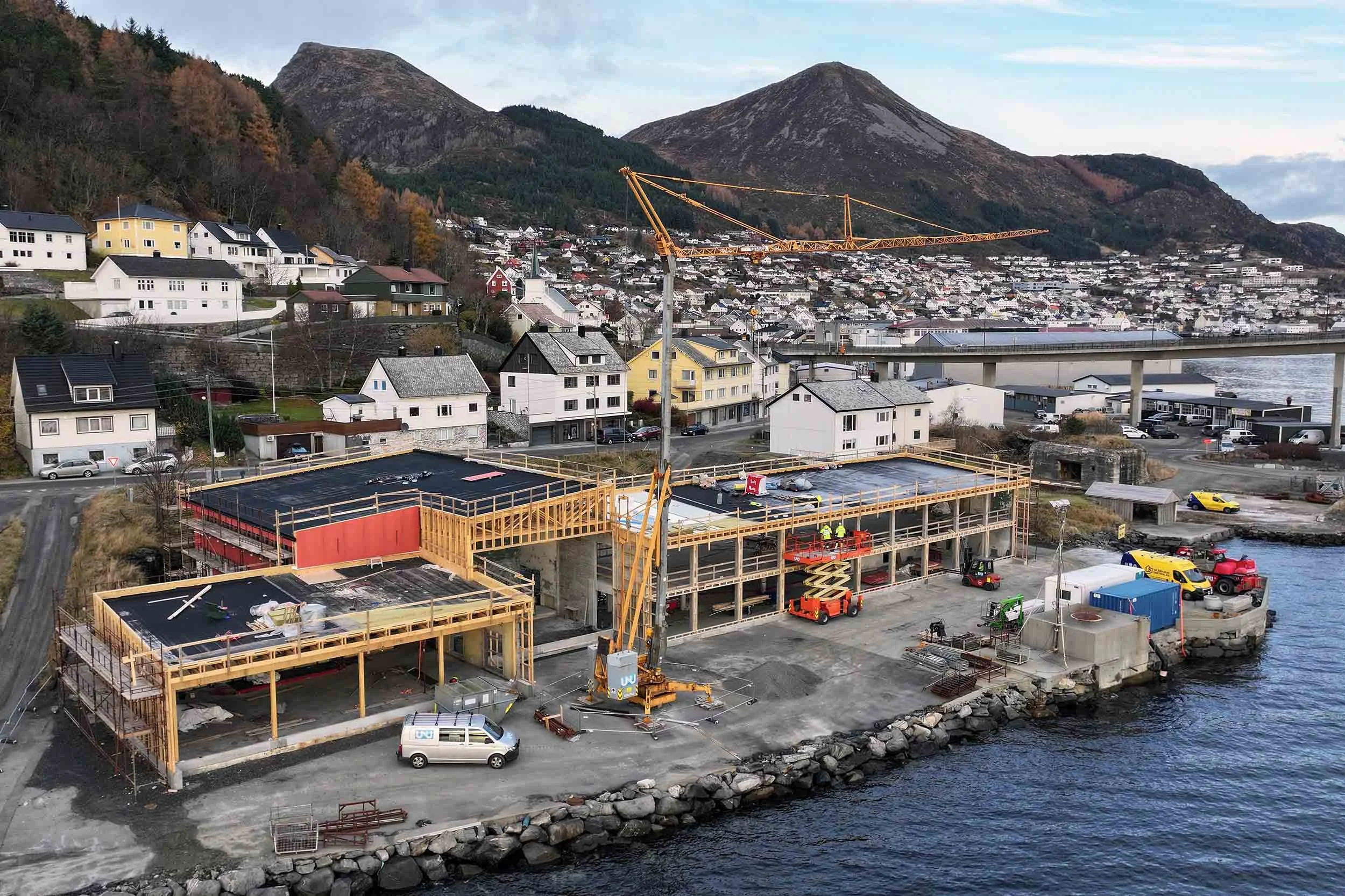Bygging av et nytt byggverk ved sjøen med et kran og byggemateriell, omgitt av hus og fjell i bakgrunnen.