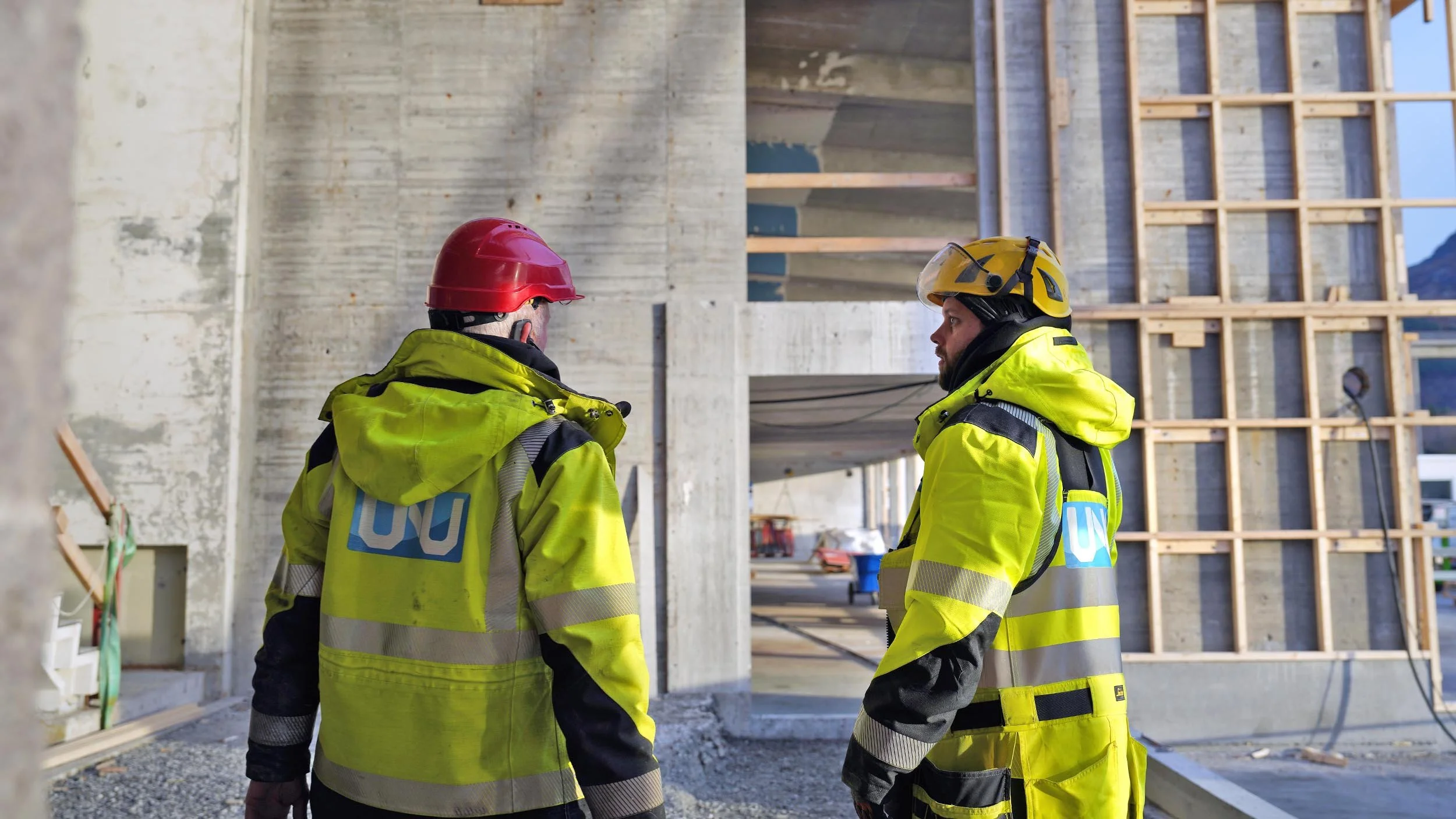 To bygningsarbeidere i gule sikkerhetsjakker og hjelmer står og snakker på en byggeplass.