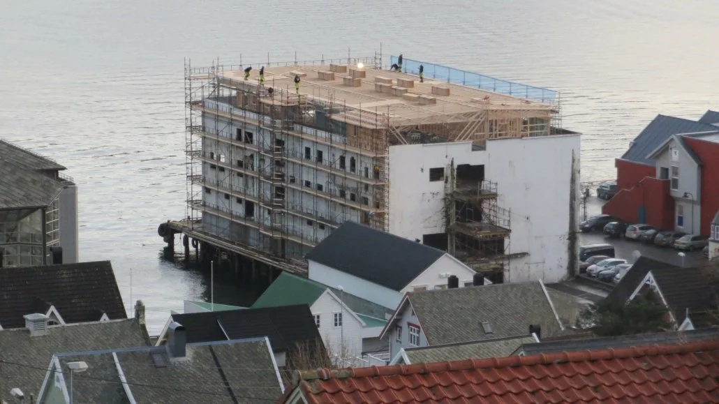 Et byggverk under konstruksjon over vann, omgitt av hus, med scaffolding og arbeidere på toppen.
