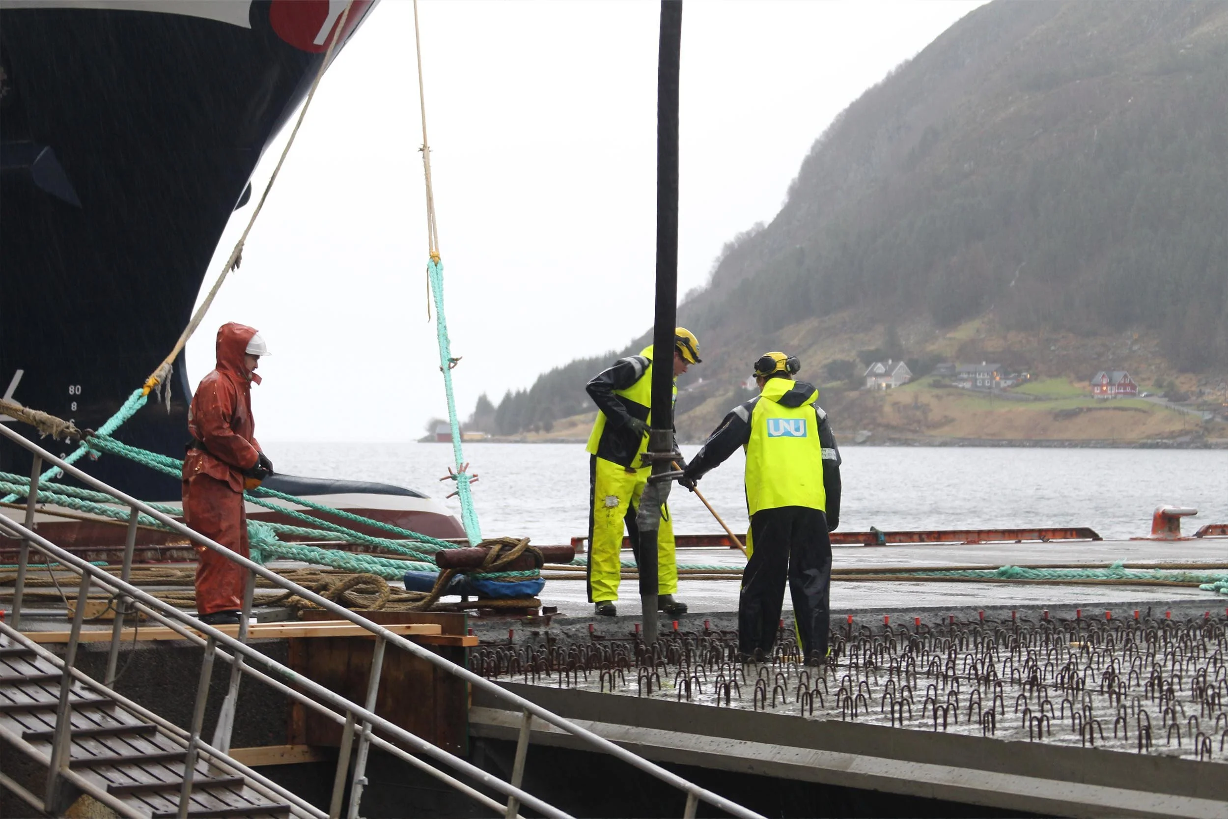 To personer i høye synsjakker jobber på en flytebrygge, med et skip i nærheten, og et fjell i bakgrunnen.