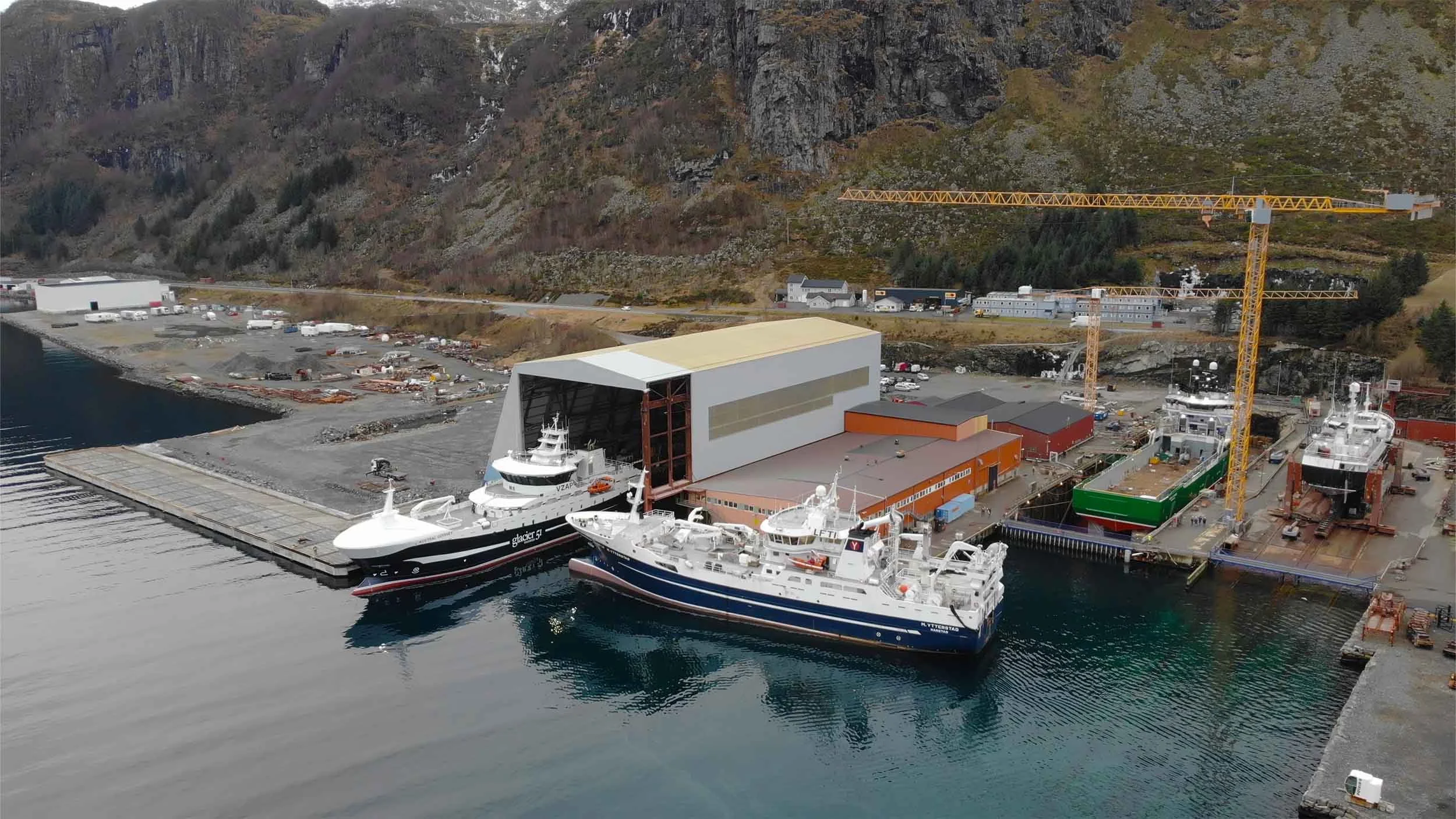 Bilde av en havn med flere båter, en stor kran og industrielle bygninger, omgitt av fjell.