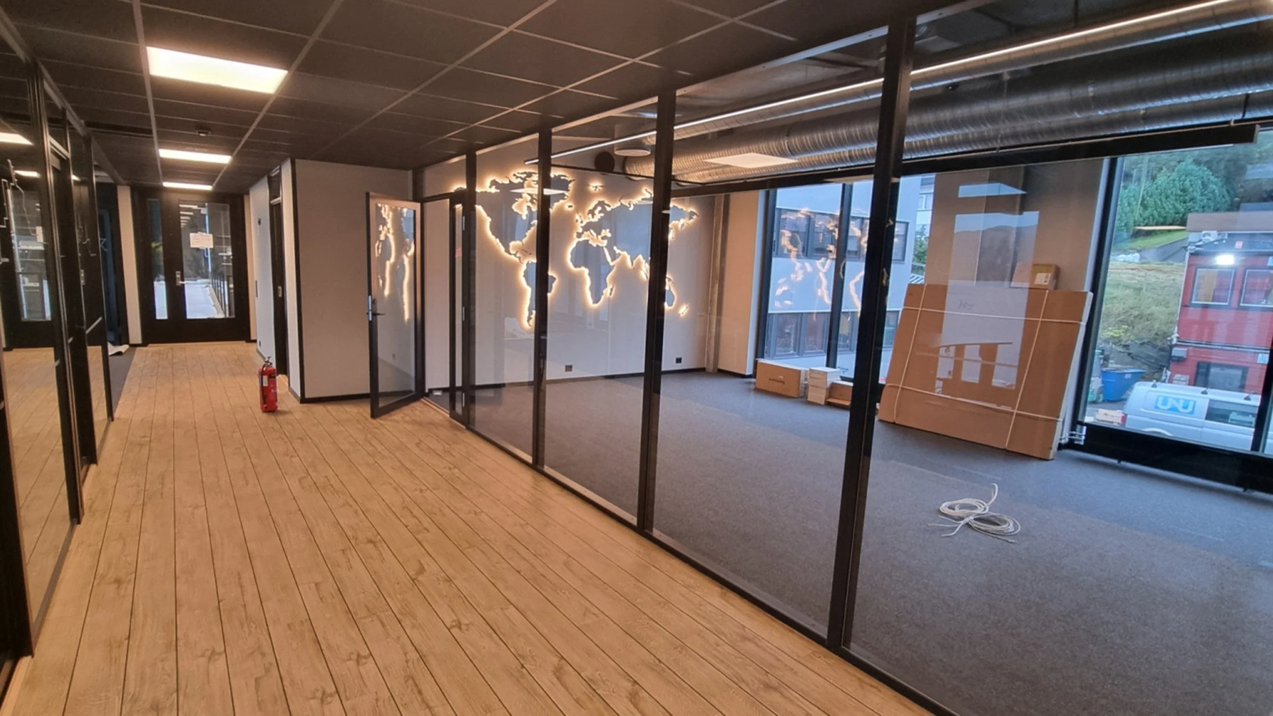 Innendørs kontor med en veggdekor som viser verdenskartet med lysende kontur. Det er glassvegger og store vinduer, med pappesker og utstyr på gulvet.
