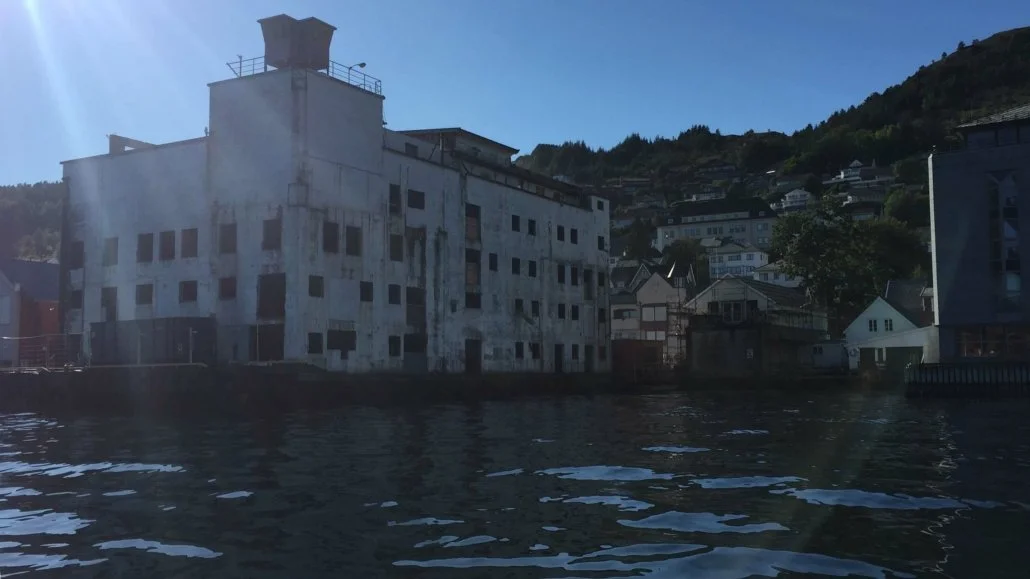 En gammel, forfallen bygning på vannet, omgitt av andre hus og fjell i bakgrunnen.