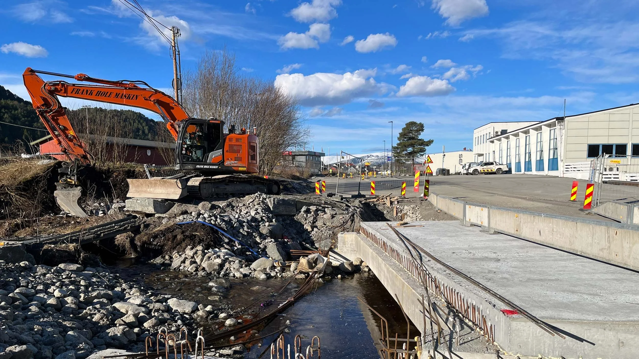 Byggearbeid med gravemaskin som fjerner bro fra en elv i en småby, med byggemeldingsskilt og noen bygninger i bakgrunnen.