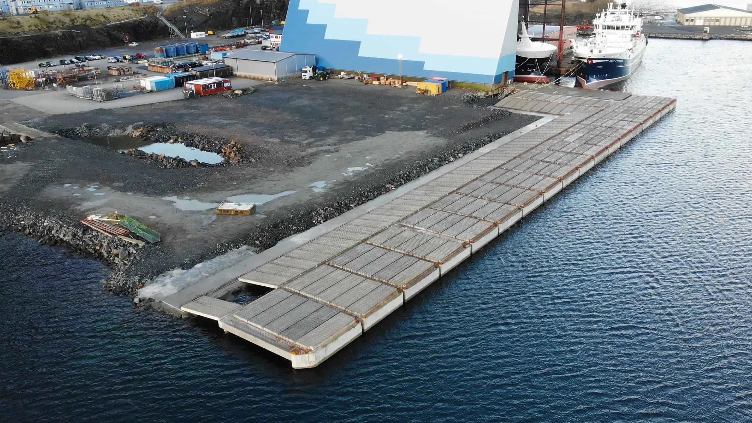 Luftbilde av en flytebrygge ved vann med to cruiseskip, en byggeplass med byggemateriell og utstyr, og en stor, blå bygning i bakgrunnen.