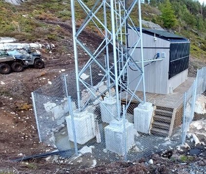 Fjernsynstårn eller transmisjonstårn med beskyttelsesgjerde rundt, plassert på en betongplattform utenfor en bygning.