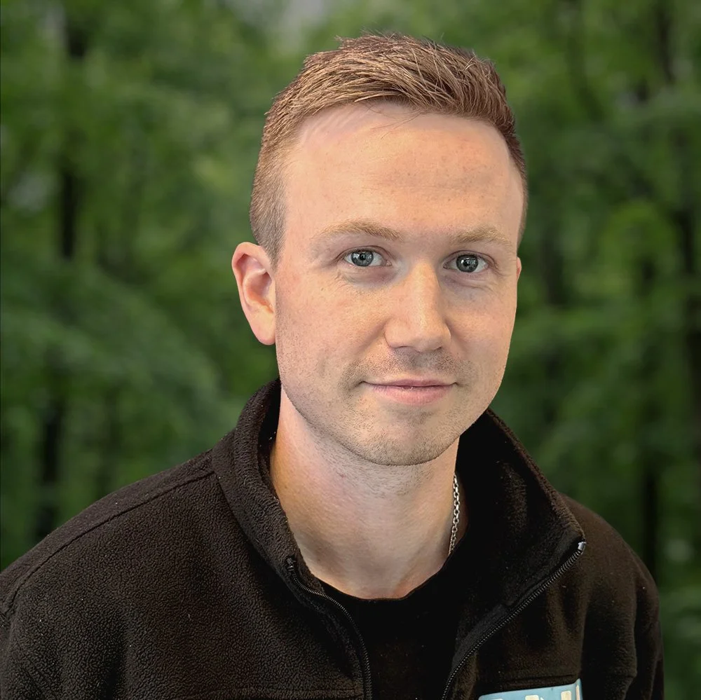 En mann med lys hud og kort, lyst hår smiler lett mot kameraet. Han har på seg en svart jakke og en kjede, med en grønn skog i bakgrunnen.