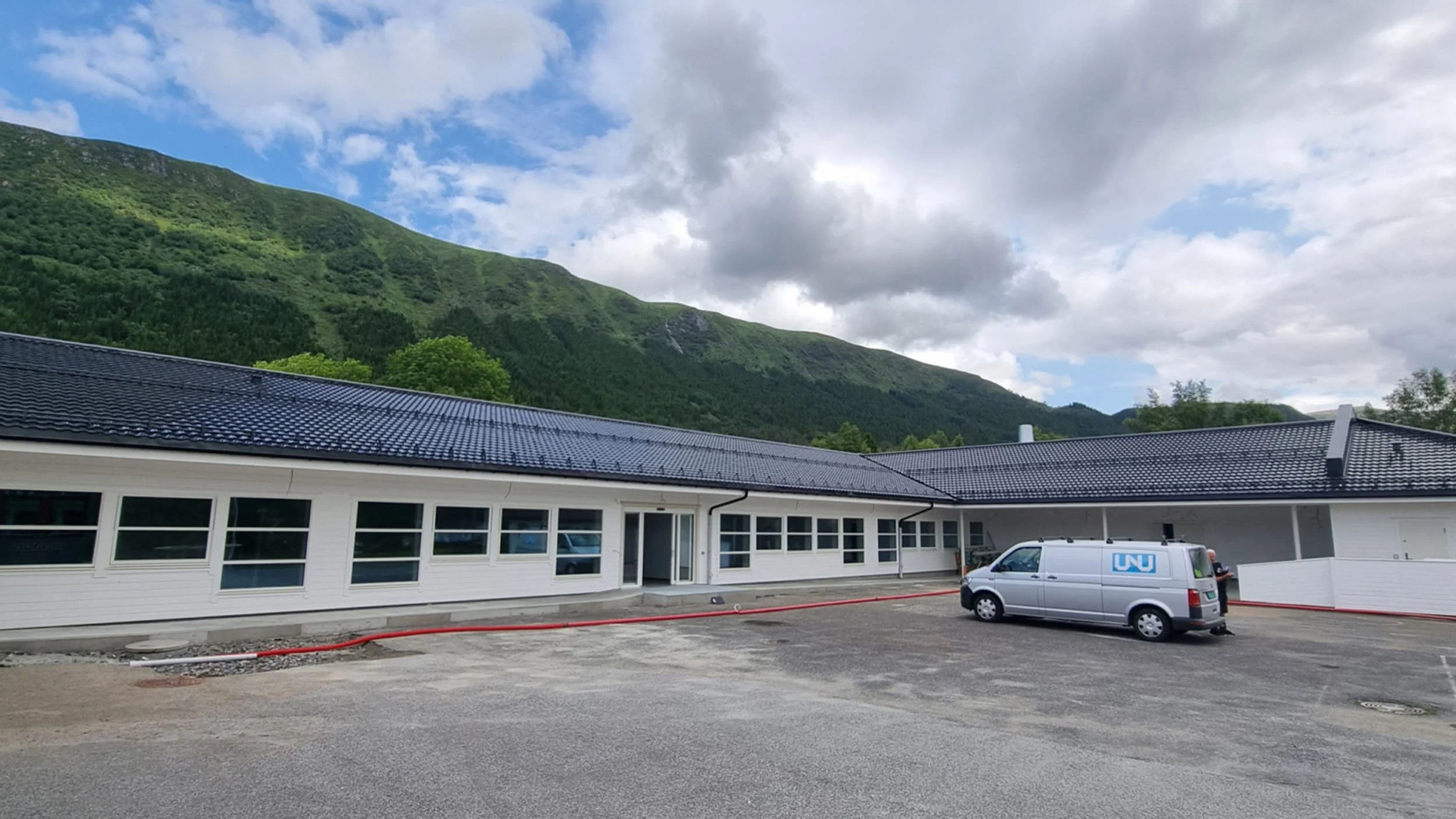 Et utendørs område med en hvit bygning med store vinduer og en parkeringsplass hvor en varebil er parkert, med grønne fjell og en blå himmel med skyer i bakgrunnen.