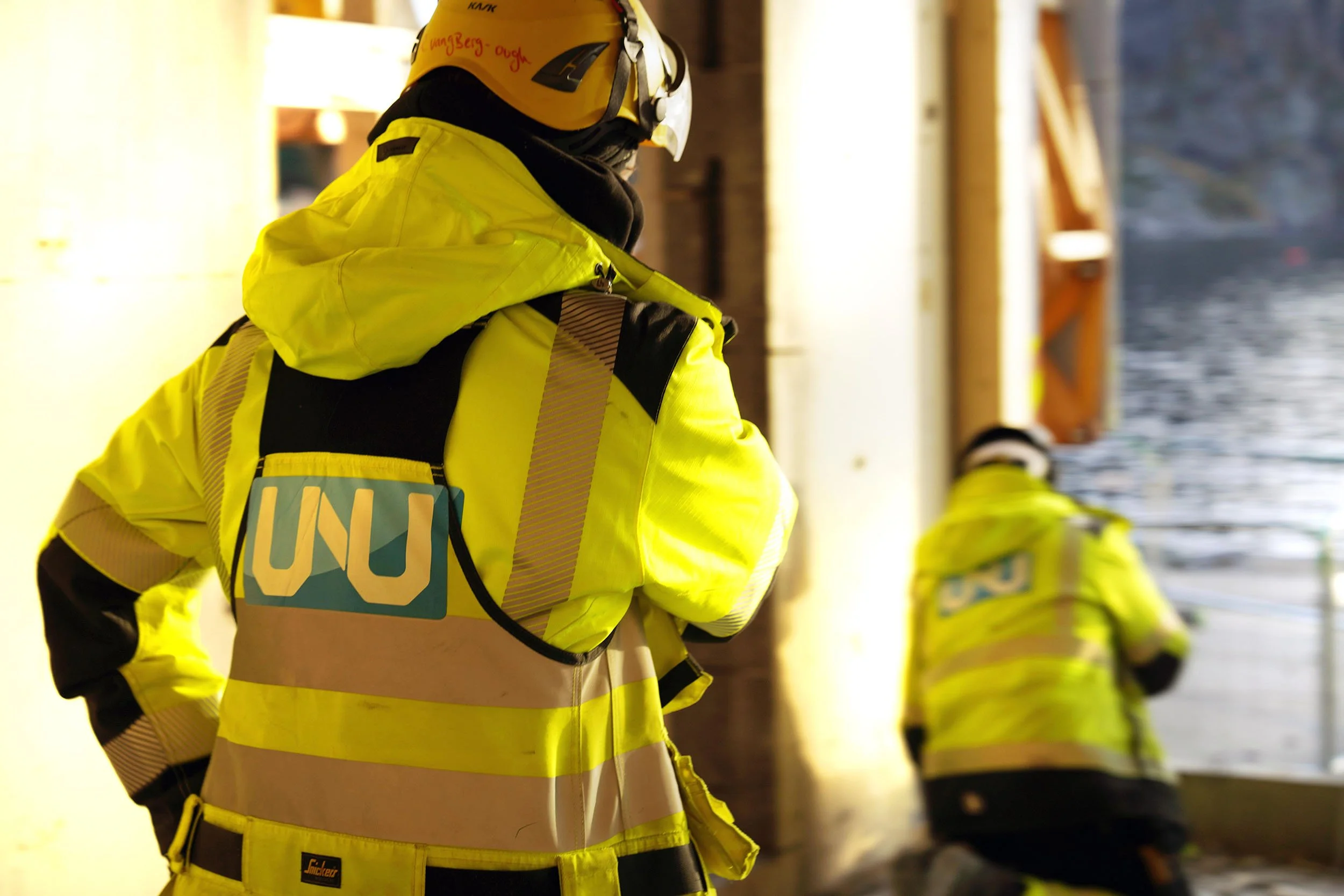 En person iført høy synlighetsjakke og hjelm, med UNODC-badge, som står i en bygning med noen andre personer i lignende klær. Personene ser ut til å være involvert i en nødsituasjon eller redningsarbeid.