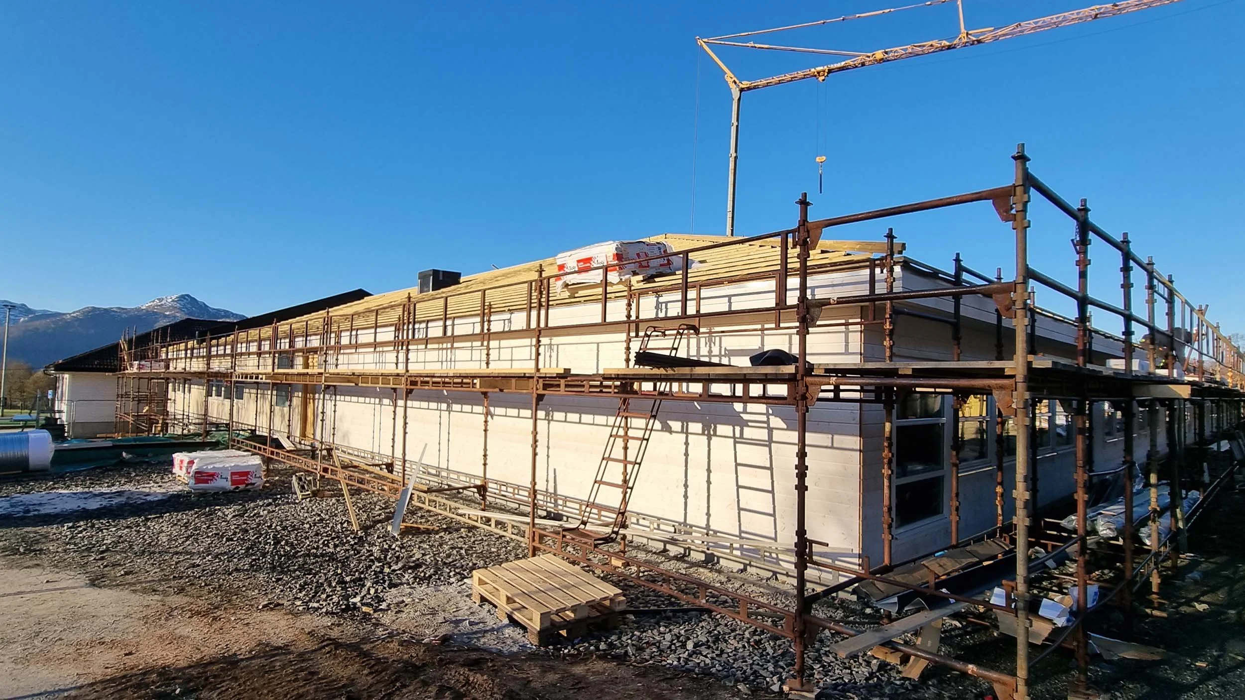 Byggefase av en moderne bolig under konstruksjon, omgitt av stillas og byggeutstyr, med fjell i bakgrunnen på klar himmel.