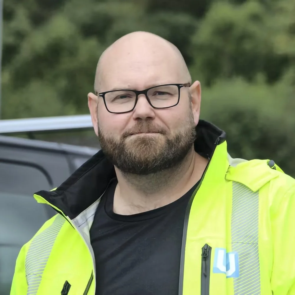 En mann med skjegg, briller og bar hode, iført en høyvis kostyme, står foran en bil med grønt landskap i bakgrunnen.