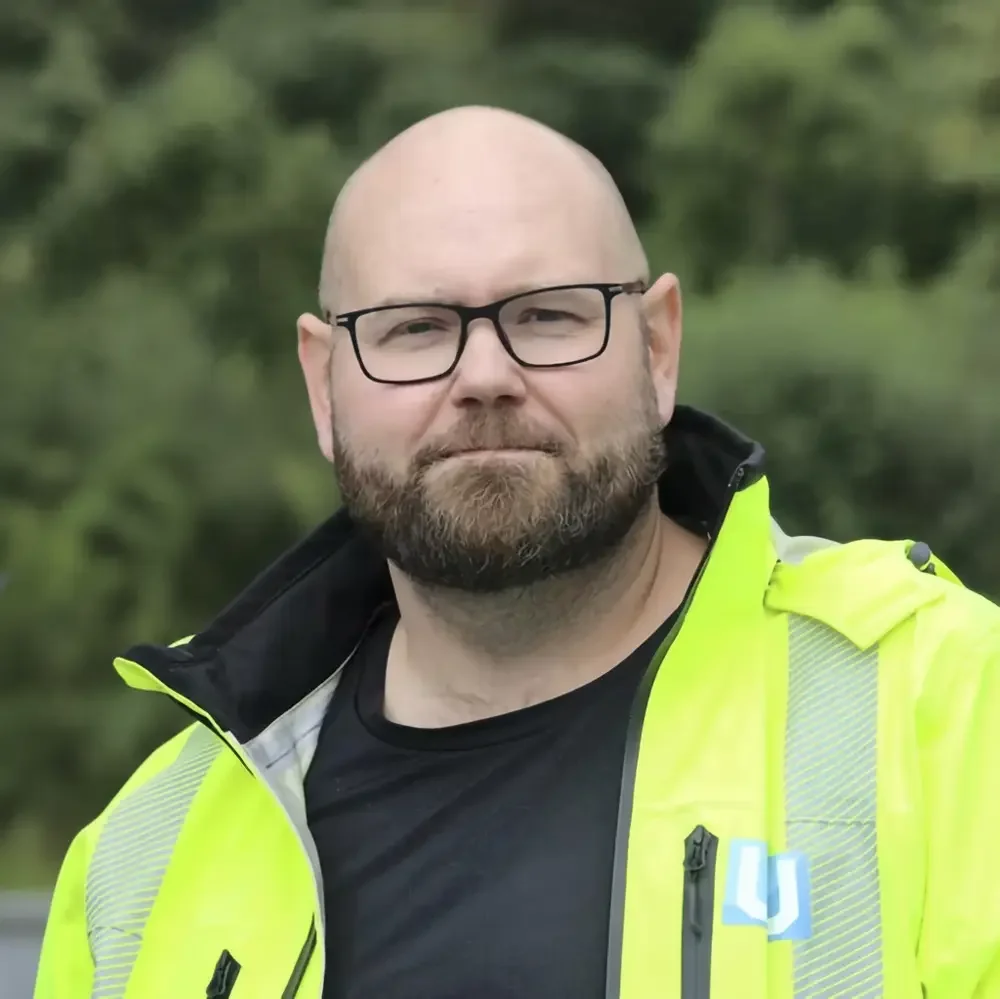 En mann med brunt skjegg og briller, iført en gul Refleksjakke, står foran en grønn bakgrunn.