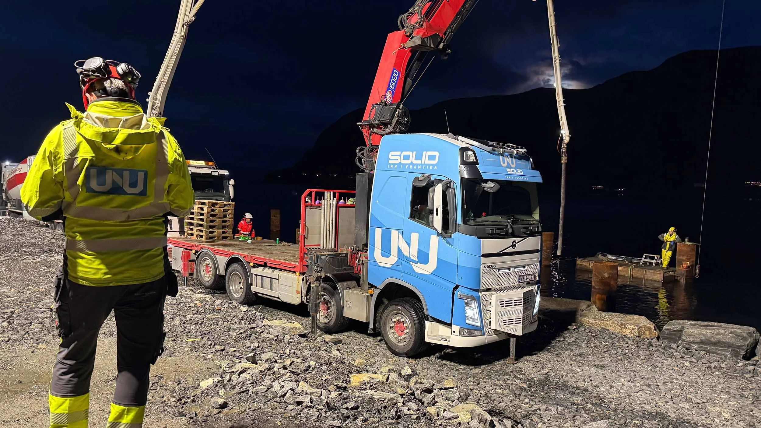 Nattbilde av redningsarbeidere i høye synsjakker og hjelmer ved en vannkropp, med redningsutstyr og en truck med en kran, under mørke himmelbakter og fjell i bakgrunnen.