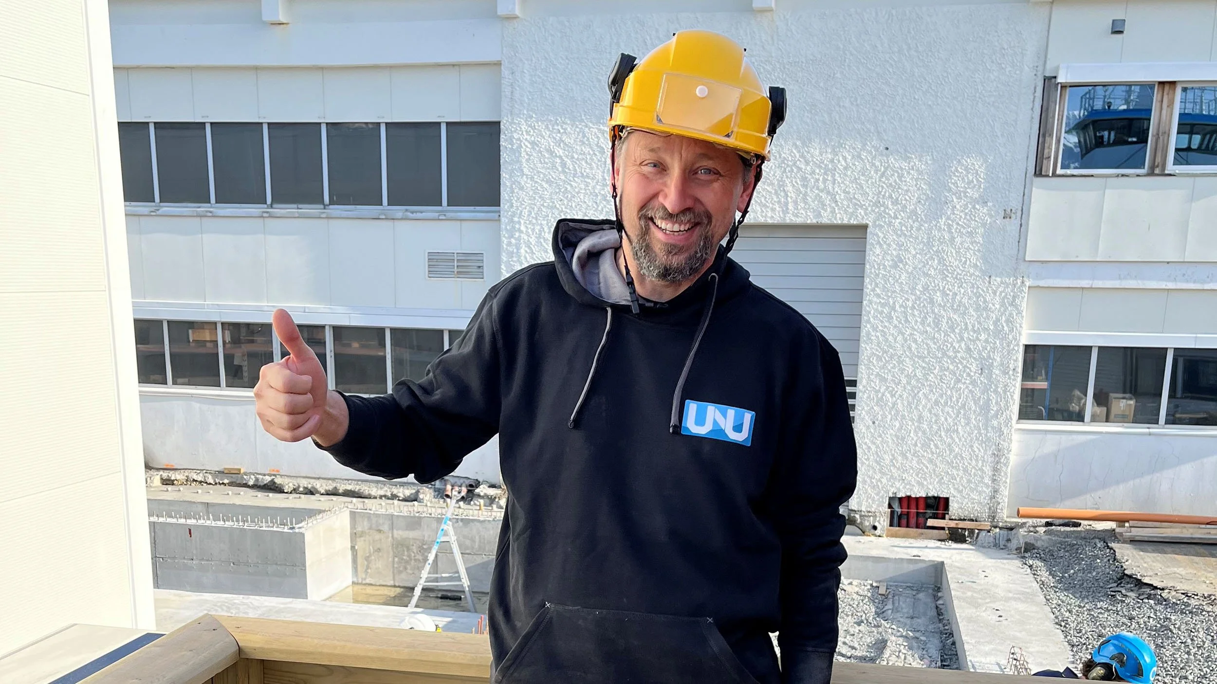 En mann med skjegg og smilende ansikt, iført gul hjelm og svart jakke med UNU-logo, står på en byggeplass med en hvit bygning i bakgrunnen og gir tommelen opp.