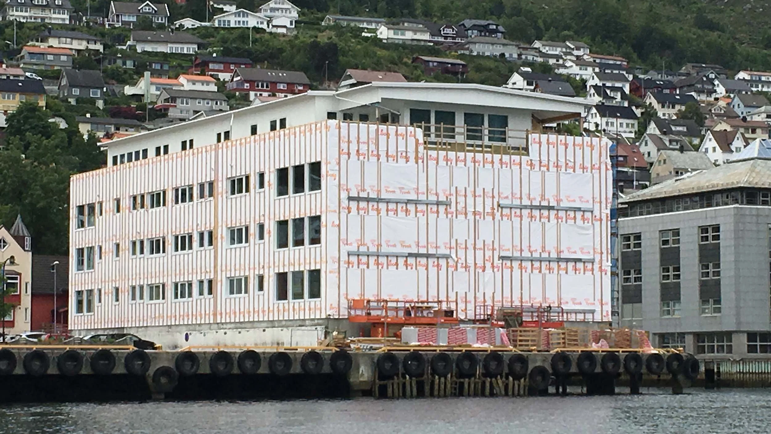 Påbegynt bygging av en gråhvitt bygning med mange vinduer, flere etasjer, og et bygg under konstruksjon i forkant. Bygningen ligger ved vannet, med en bygd av hus i fjellet i bakgrunnen.