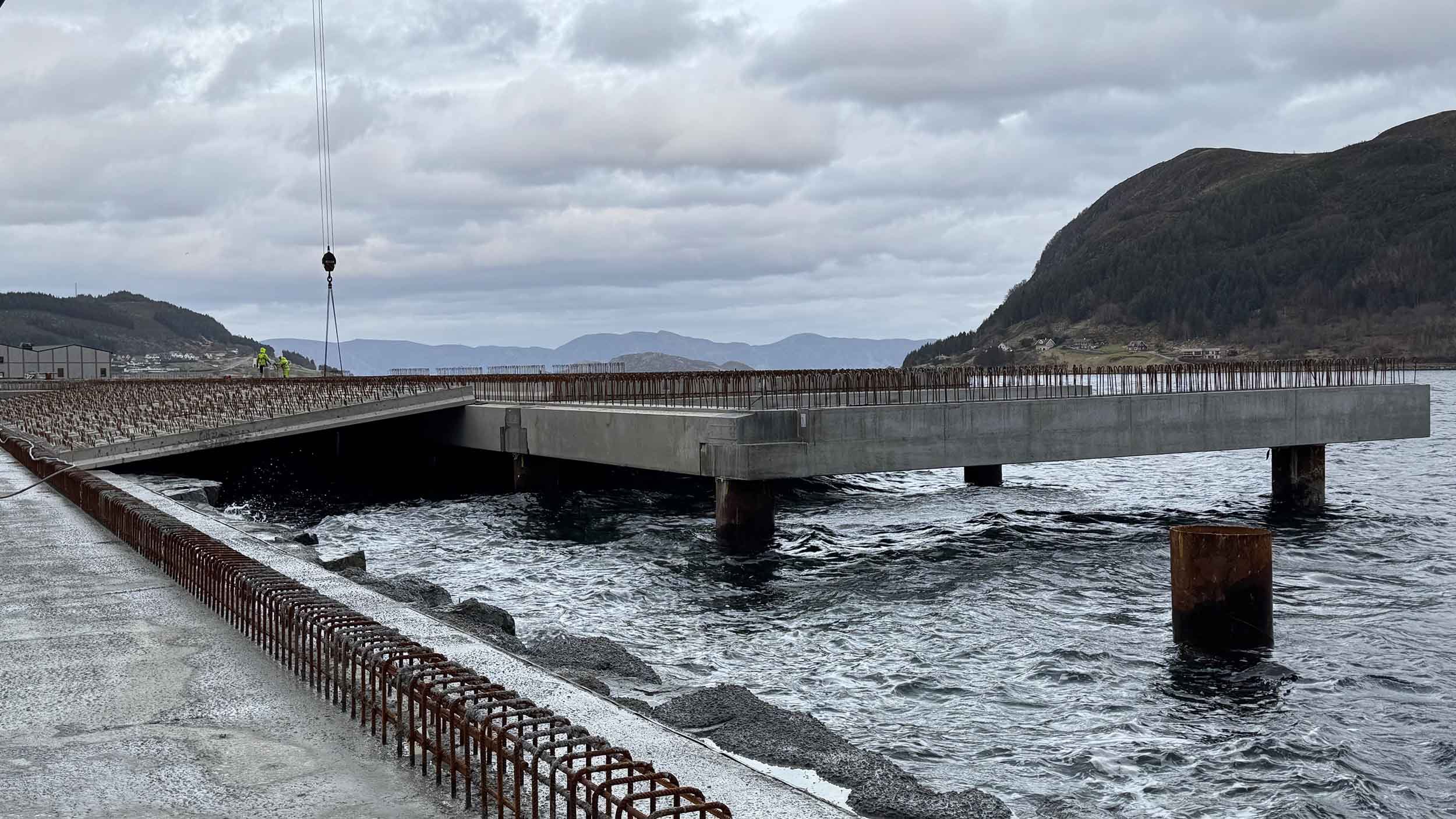 På byggeplass med en ny bro under konstruksjon ved vann, fjell i bakgrunnen og to arbeidere i lyse arbeidsklær.