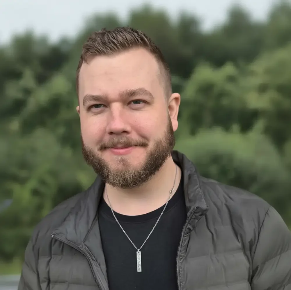 Profilbilde av en mann med kort brun hår, skjegg, svart genser og jakke, mot en grønn bakgrunn.