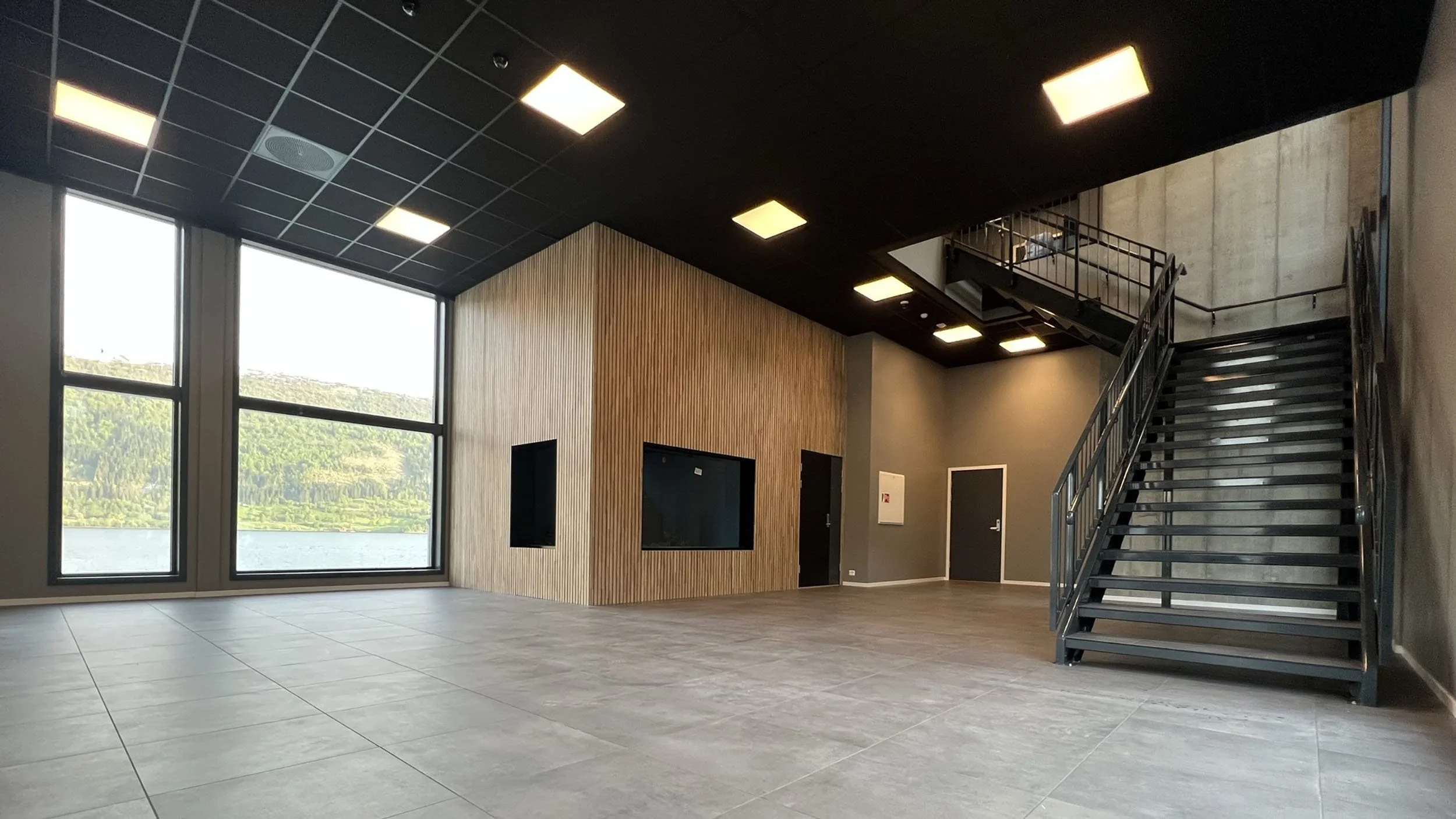 Innendørsområde med store vinduer, trevegg og metalltrapp.