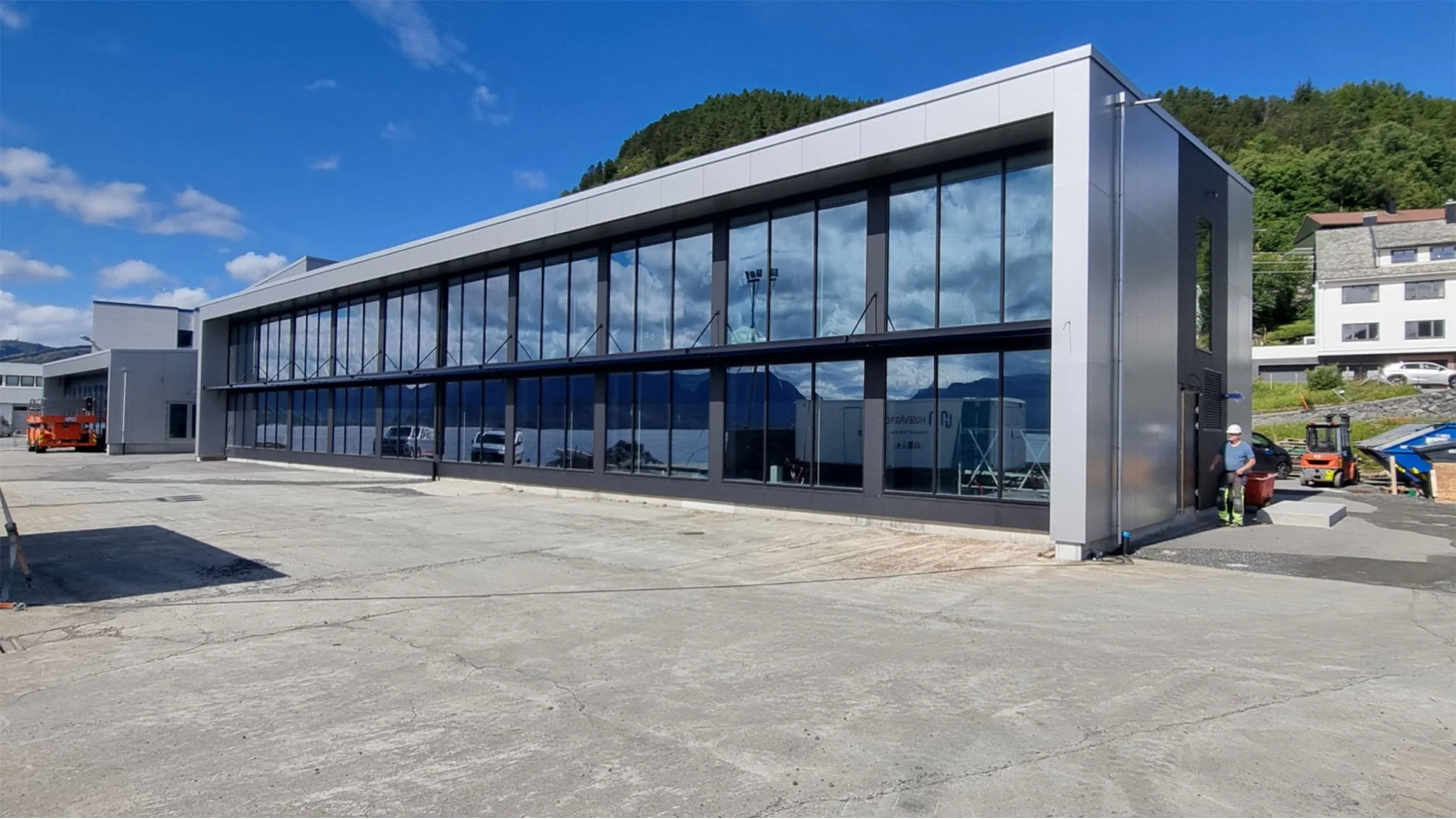 Nytt moderne kontorbygg under bygging, store glassvinduer og byggemøbler rundt, flere bygninger i bakgrunnen, vakker fjellandskap og blå himmel.