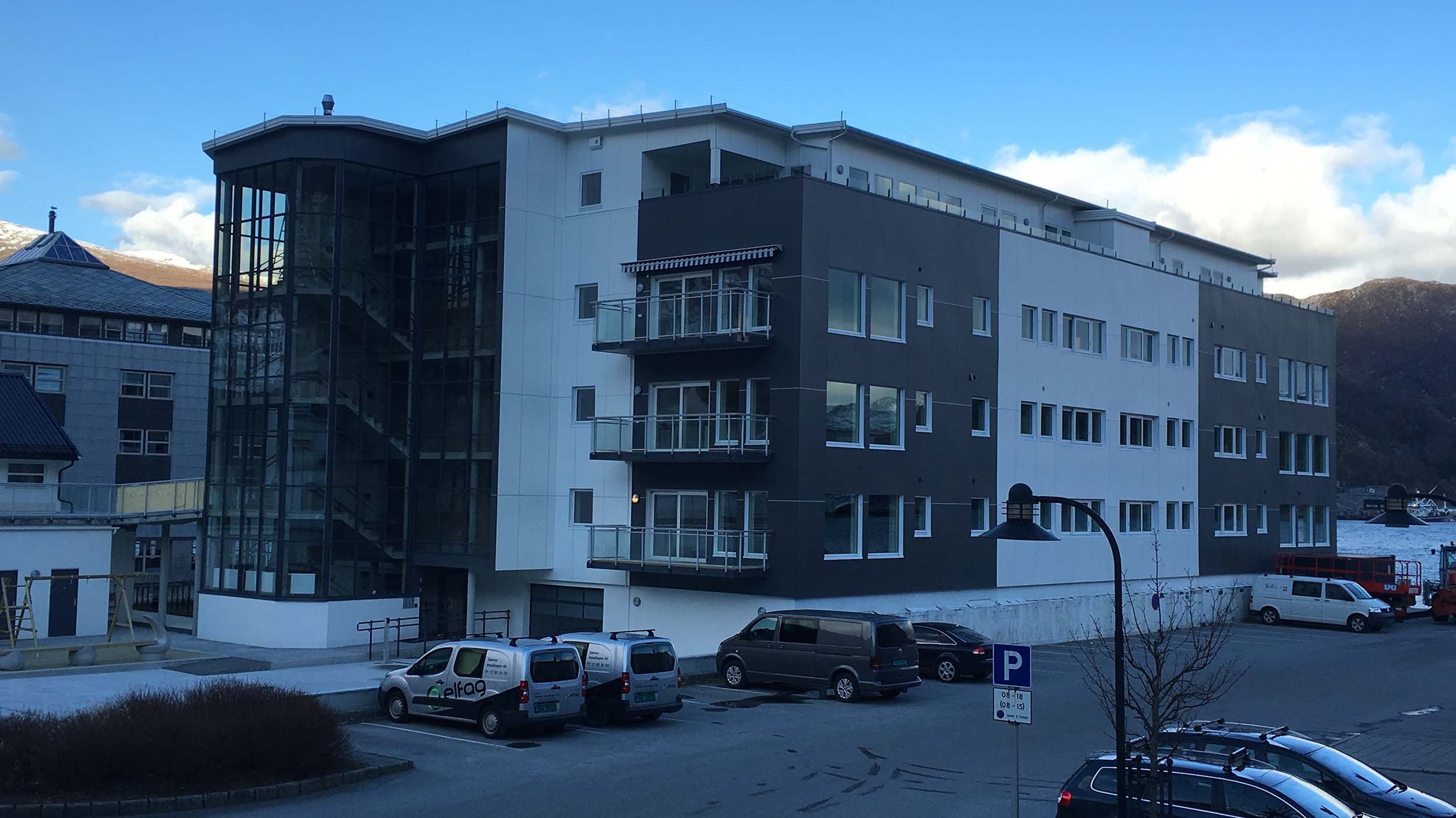 Moderne, fire-etasjers boligbygg med store vinduer og balkonger, omgitt av biler i en parkeringslade, med fjell i bakgrunnen og en klar blå himmel.